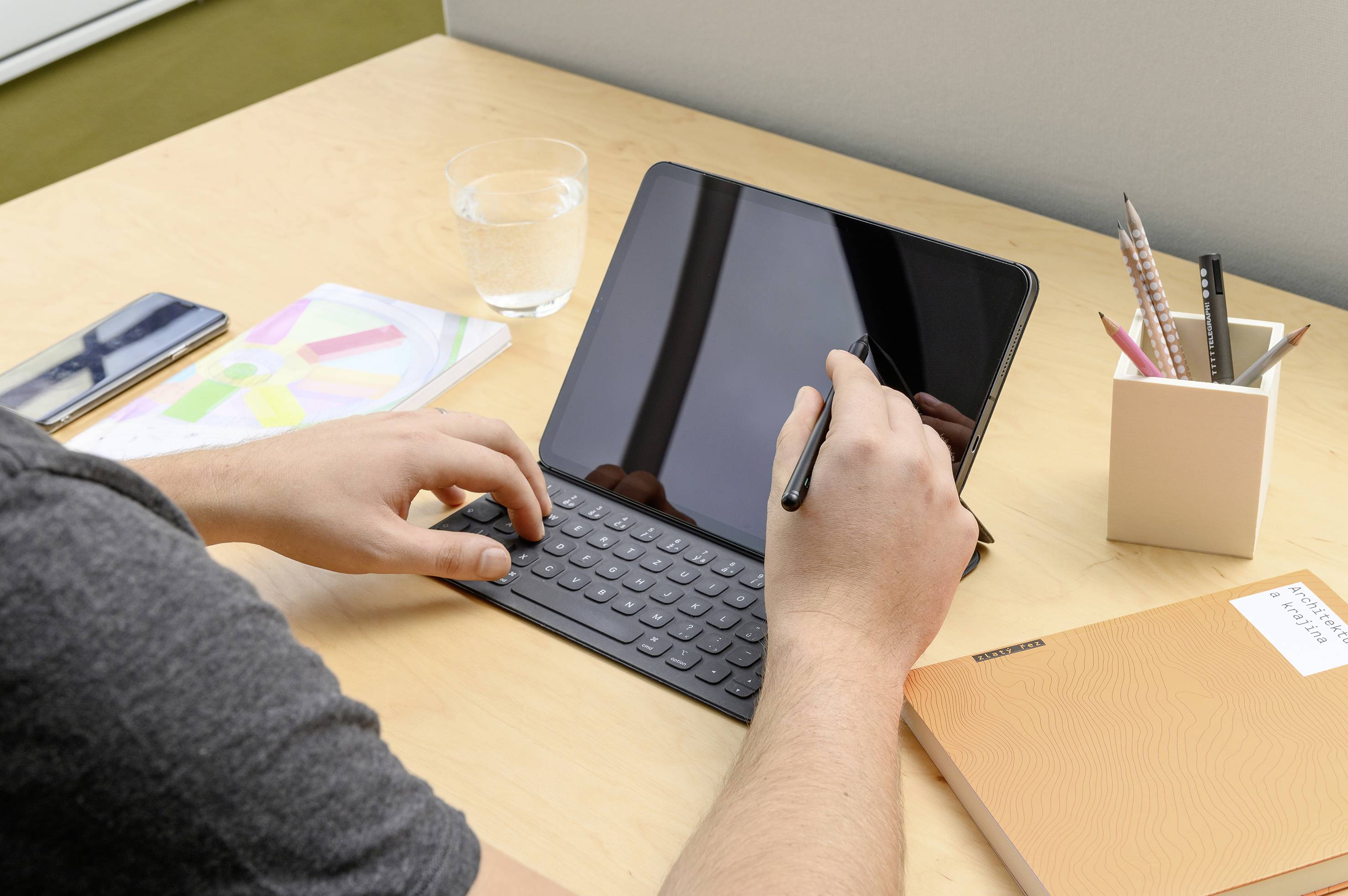 Osoba pisze długopisem na tablecie z klawiaturą. Na biurku leżą notatki, długopisy, szklanka wody.