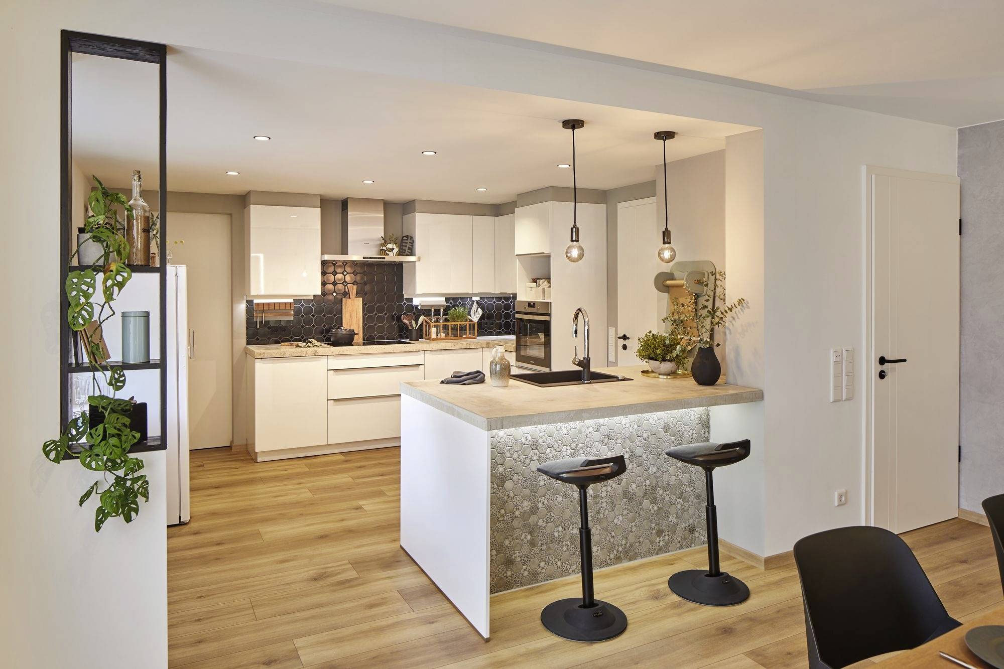 Nowoczesna kuchnia z białym wyposażeniem, drewnianą podłogą i wyspą z dwoma czarnymi stołkami barowymi. Wiszące lampy zapewniają oświetlenie.