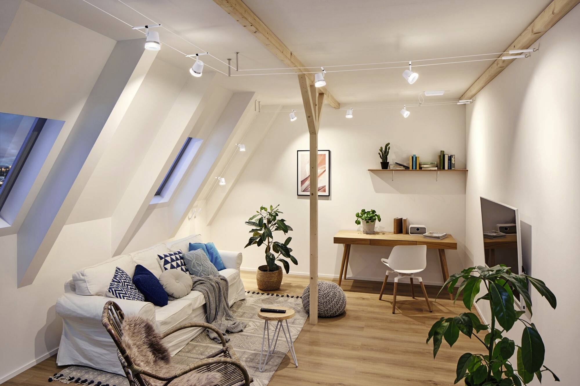 Przytulny salon z ukośnym sufitem. Kanapa z poduszkami, rośliny, biurko z krzesłem i półkami. Jasne oświetlenie.