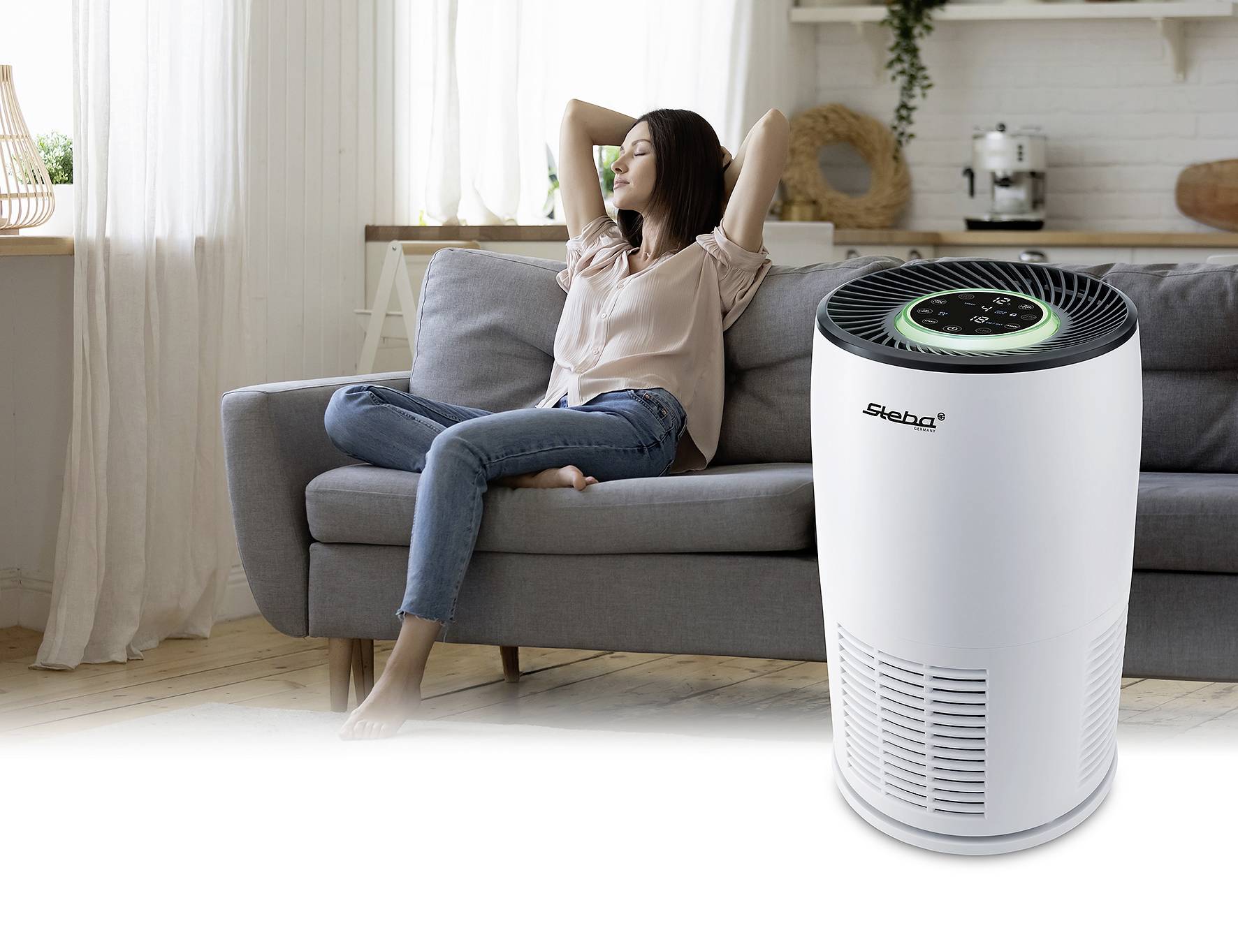 Kobieta relaksuje się na kanapie, a w pierwszym planie widoczny jest oczyszczacz powietrza z podświetlanym panelem sterowania. W tle widać wystrój salonu.