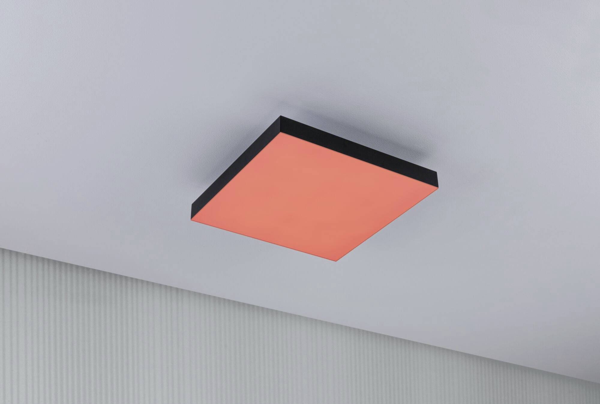Kwadratowa, płaska lampa sufitowa świeci intensywną czerwienią i kontrastuje z białym sufitem w nowocześnie urządzonym pomieszczeniu.