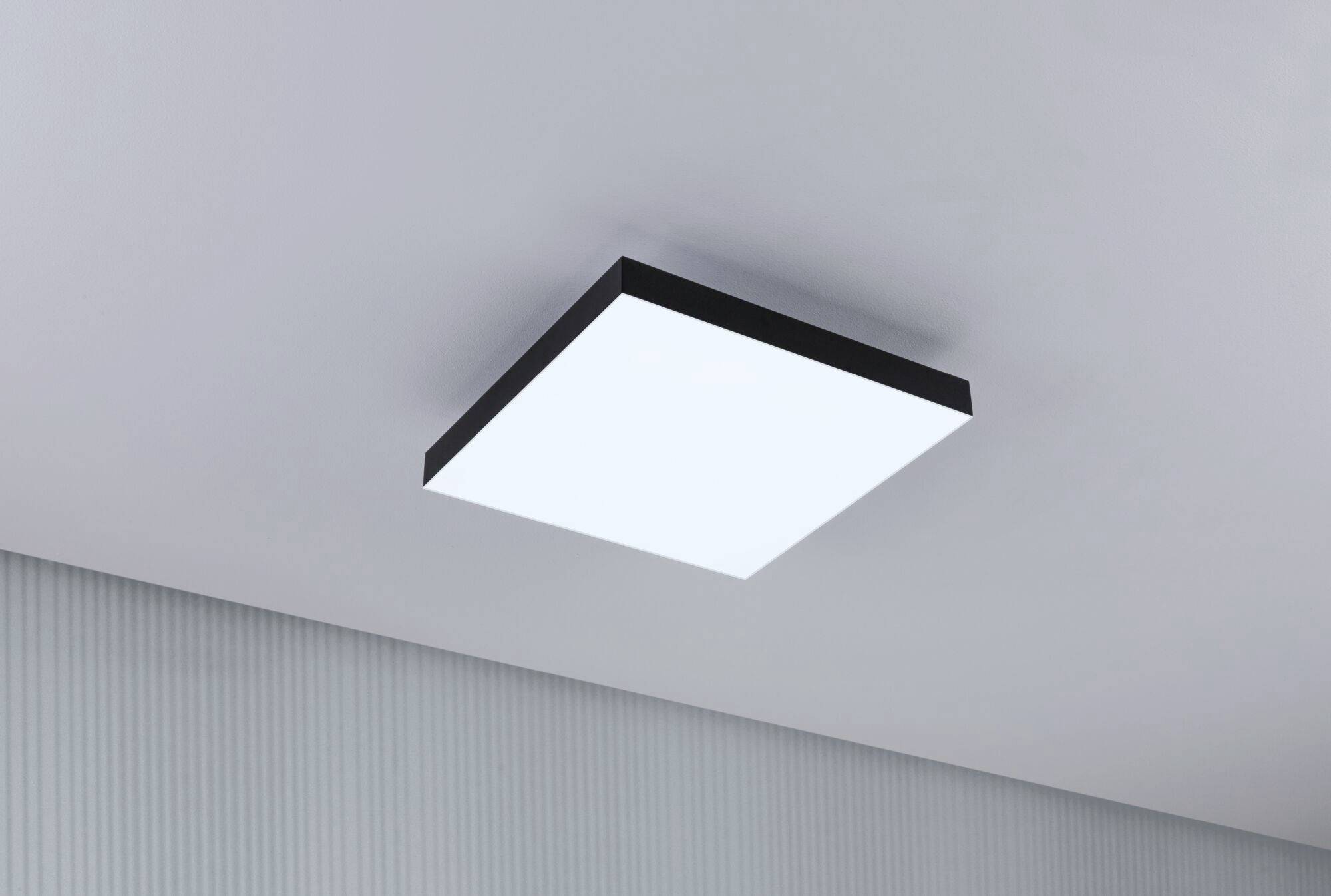 Prostokątna lampa sufitowa z prostą czarną ramką jest zamontowana na szarym suficie i świeci jasnym światłem.