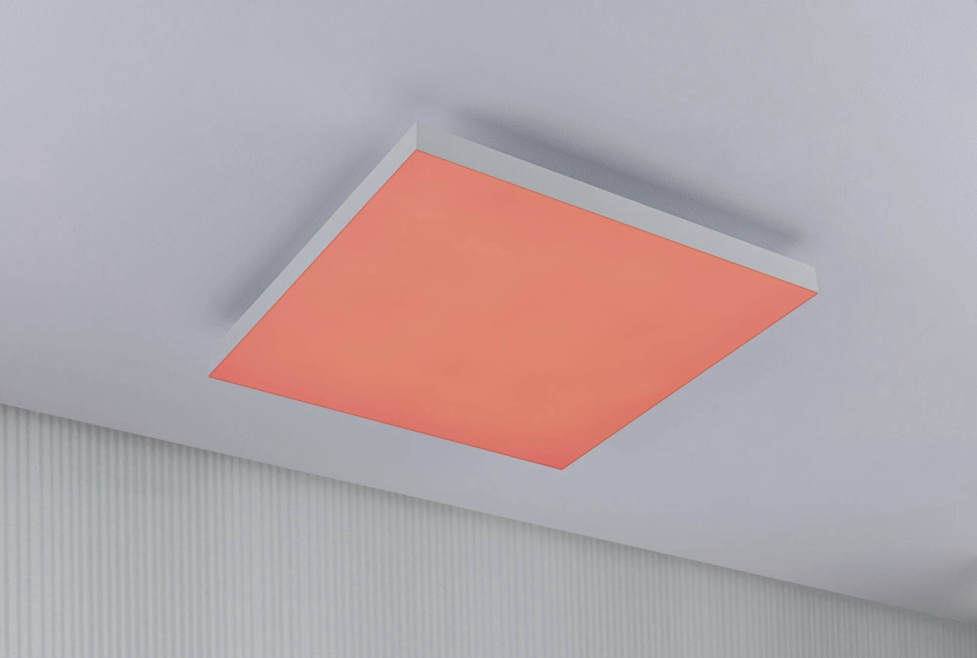 Kwadratowa, czerwona lampa sufitowa świeci w jasnym pomieszczeniu o białej, fakturowanej ścianie. Minimalistyczny design.