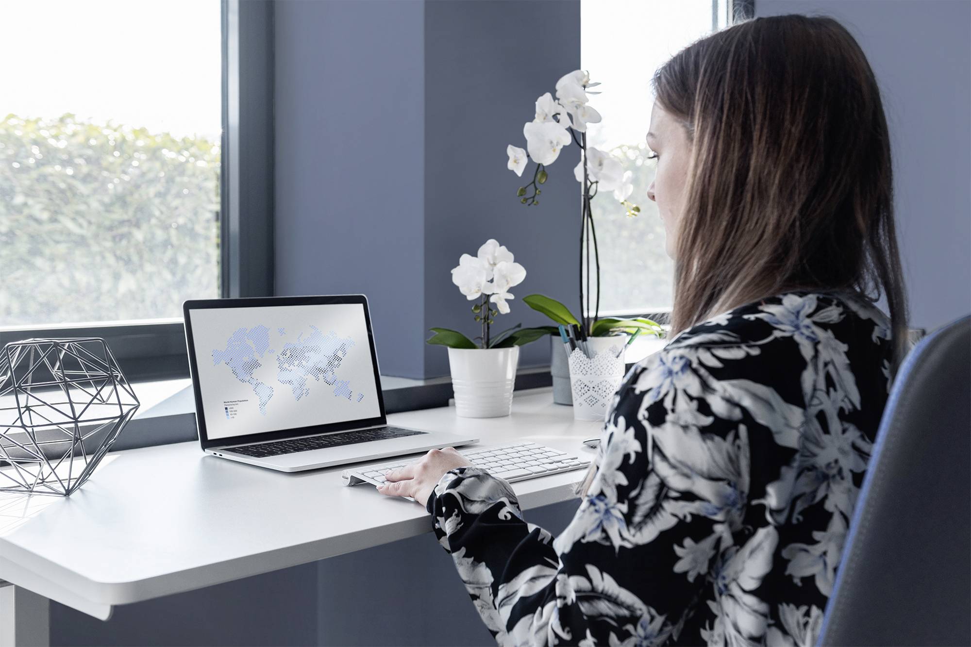 Kobieta siedzi przy stole i pracuje na laptopie. Na ekranie widać mapę świata. W tle widoczne są orchidee i geometryczne dekoracje.