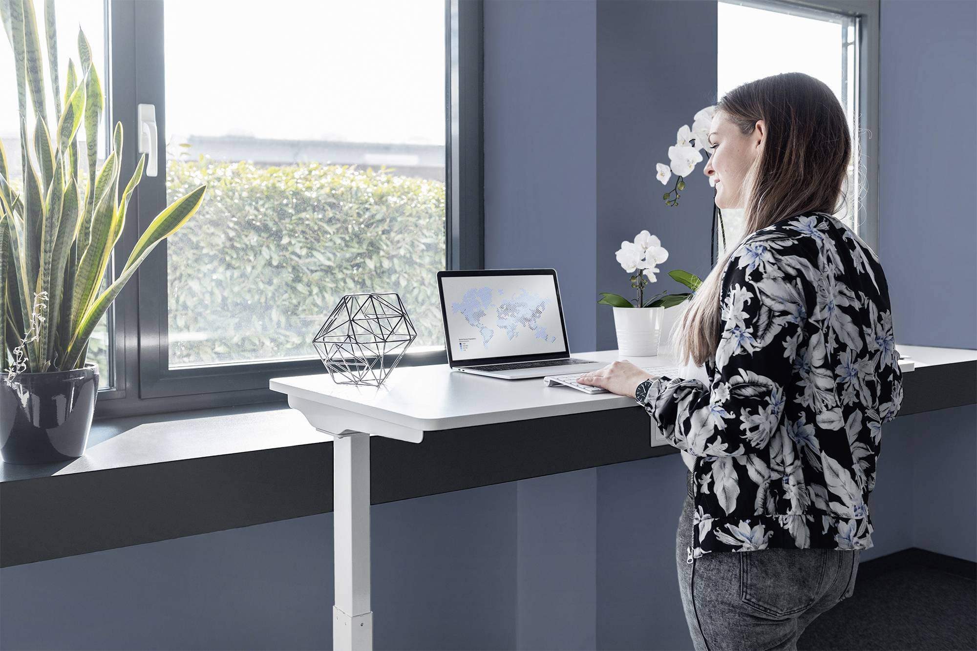 Kobieta stoi przy biurku z regulowaną wysokością i pracuje na laptopie. W tle widać rośliny i okno.