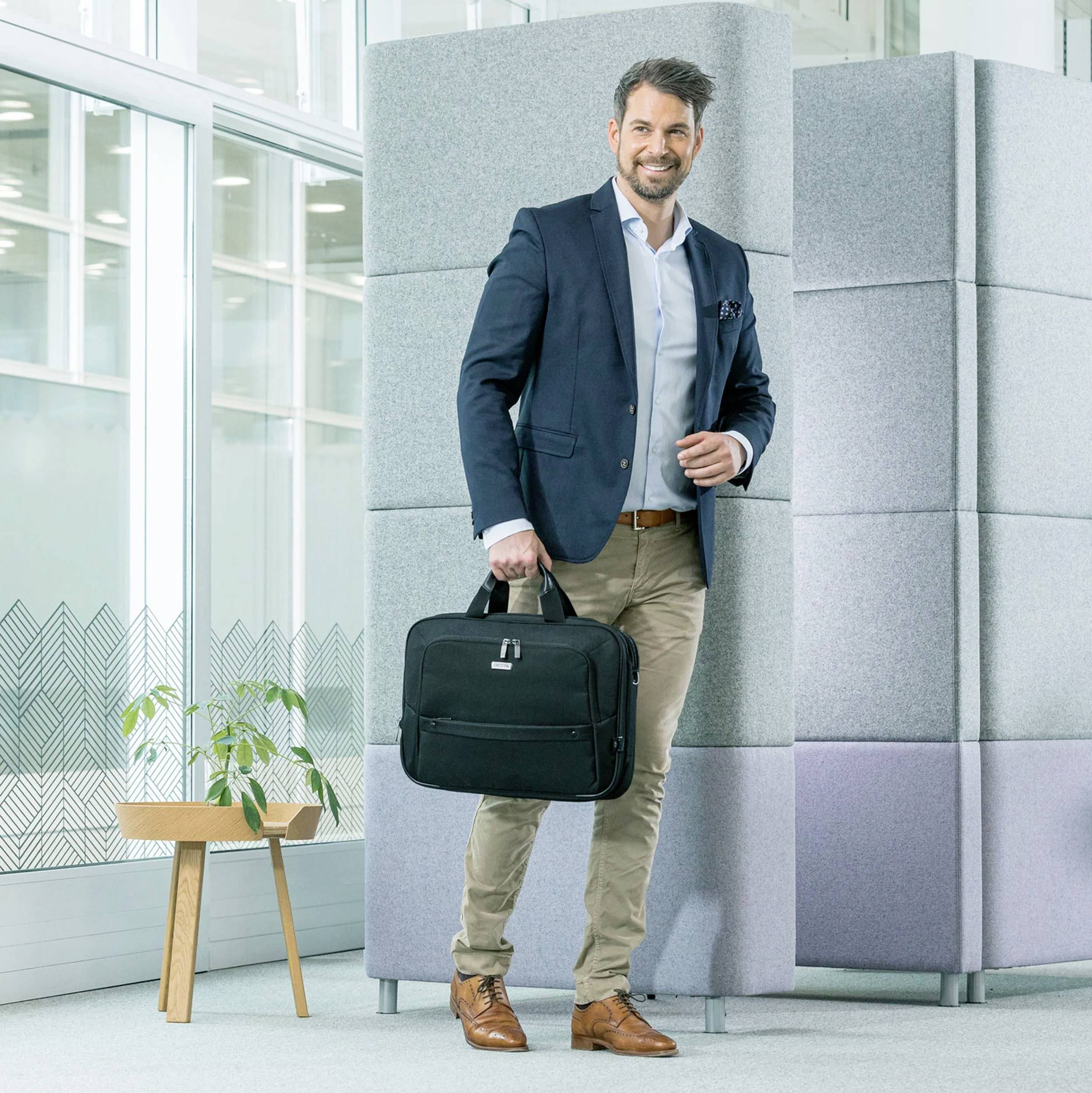 Mężczyzna w garniturze stoi uśmiechnięty obok szarej ściany, trzymając czarną torbę. W tle widać duże okna i stół z rośliną.
