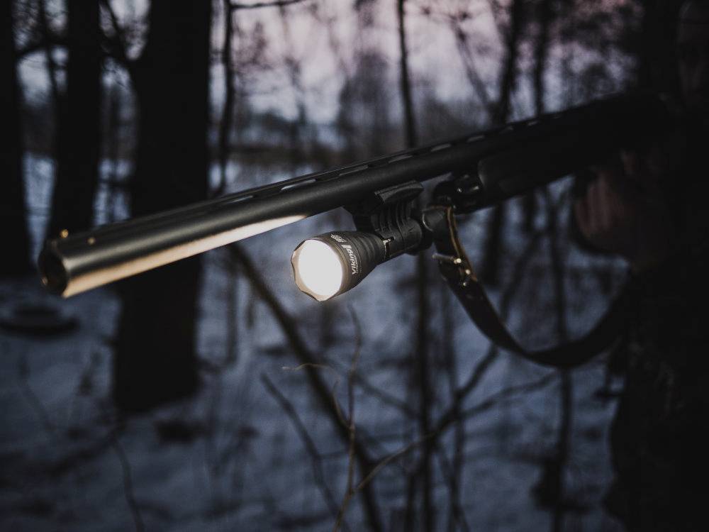 Osoba w lesie trzyma pistolet z zamontowaną latarką, skierowaną do przodu. Zmierzch zapewnia przytłumione światło.