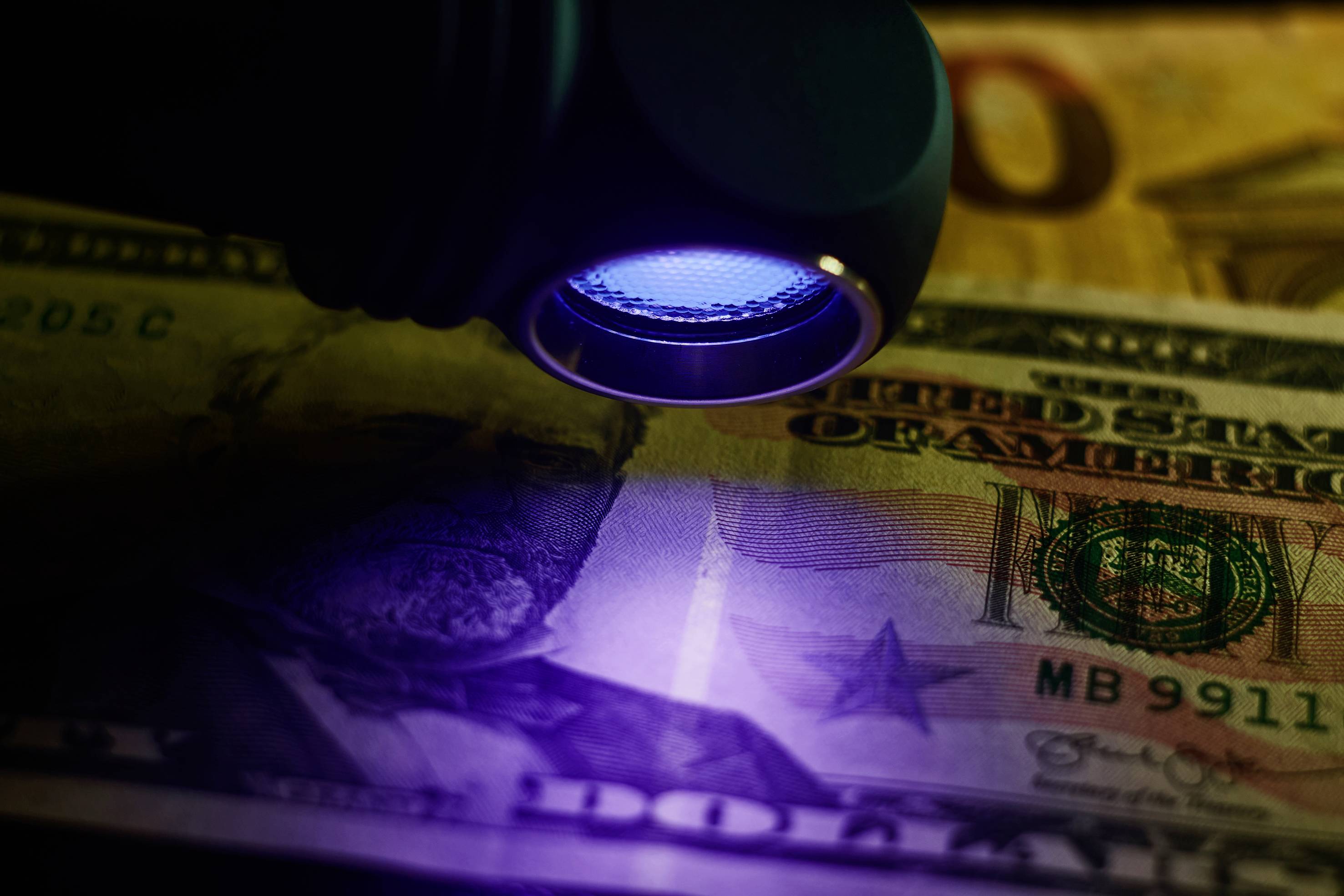 Lampa UV oświetla banknot dolara amerykańskiego. Widoczne są cechy zabezpieczające. W tle znajduje się kolejny banknot.