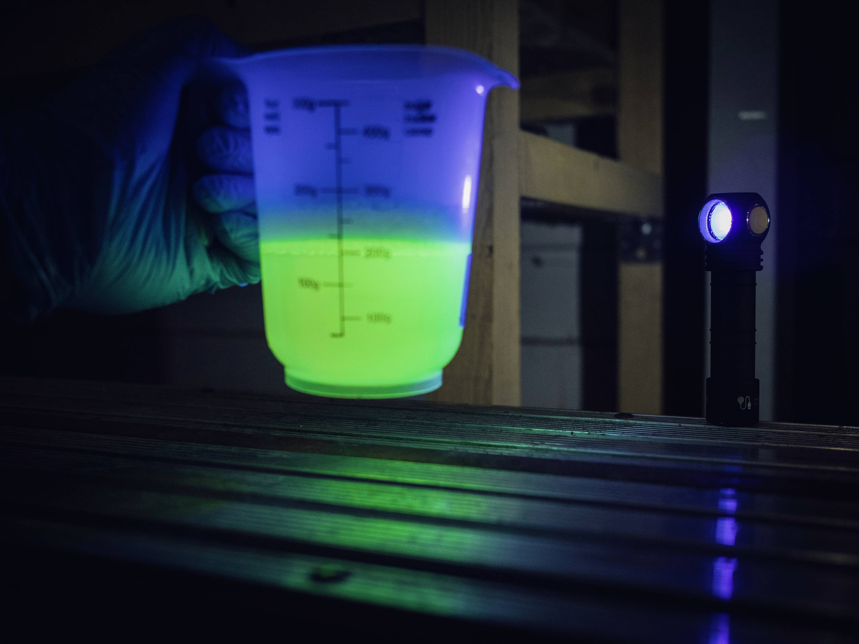 Dłoń w niebieskiej rękawiczce trzyma menzurkę z jaskrawo zieloną cieczą. Niebieska lampa oświetla scenę.