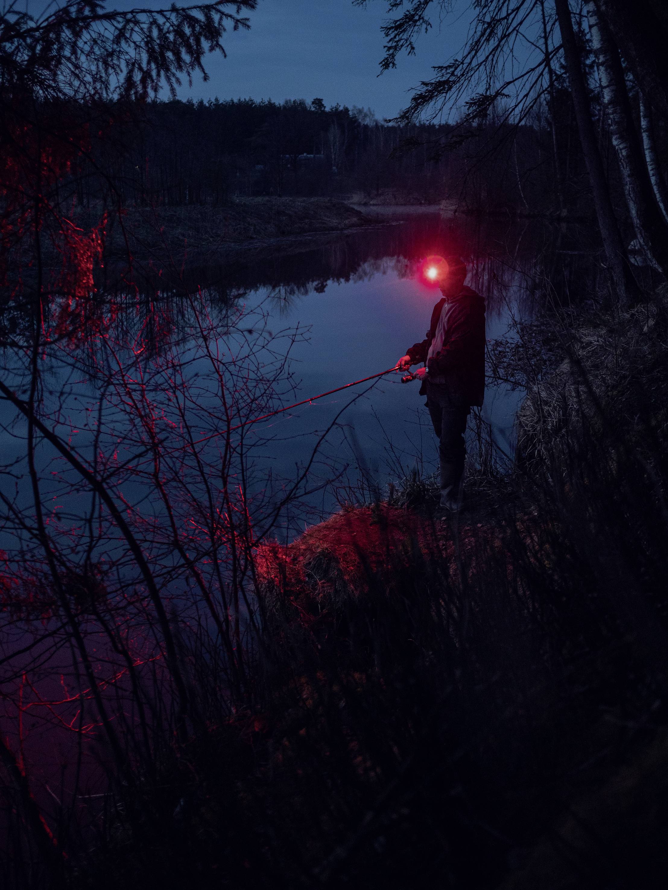 Osoba wędkuje nocą nad spokojnym jeziorem. Czerwone światło pada na wodę i otoczenie, tworząc tajemniczą atmosferę.