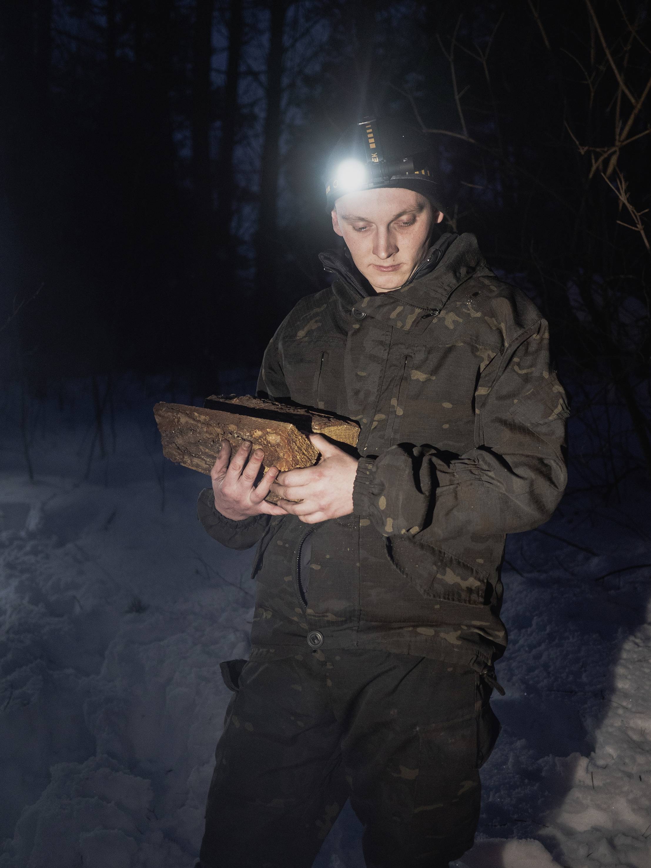 Człowiek w kamuflażu stoi nocą na śniegu i trzyma prostokątny drewniany obiekt, oświetlony czołówką.