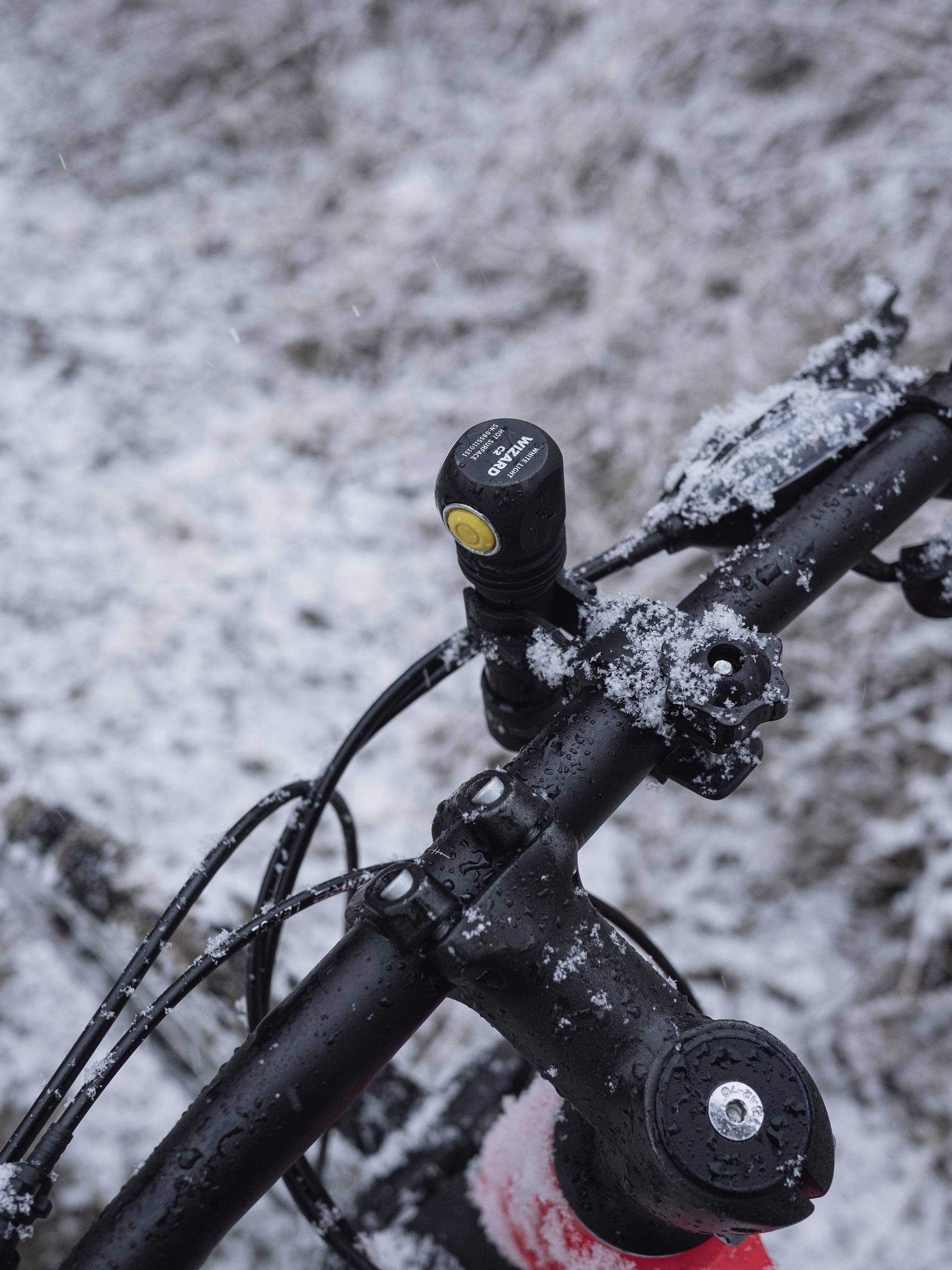 Kierownica roweru pokryta śniegiem. Mała lampka jest na niej zamontowana. W tle widać lekko padający śnieg.