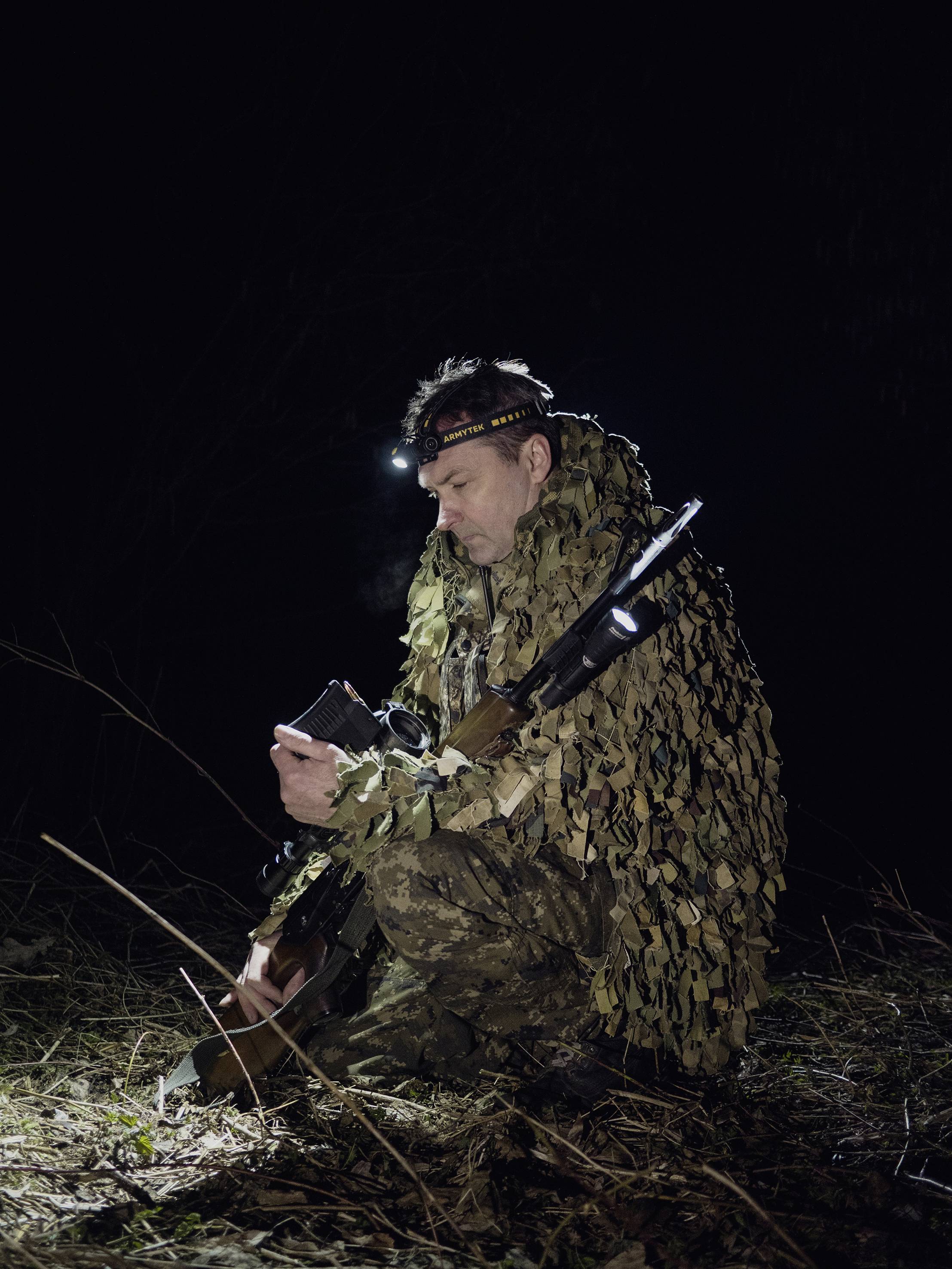 Osoba w kamuflażu siedzi nocą w lesie i patrzy na urządzenie trzymane w ręce, nosi czołówkę, obok niej leży karabin.