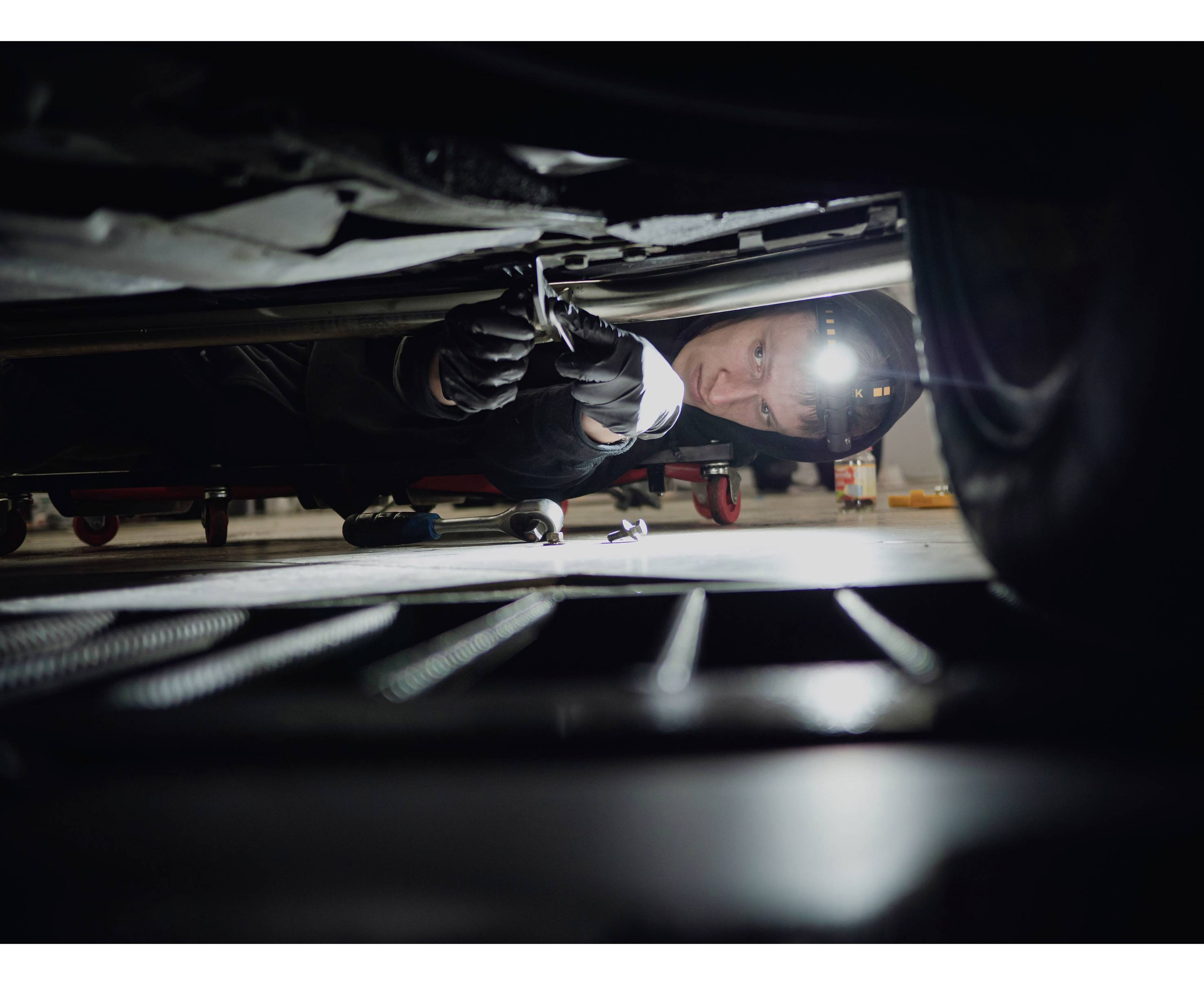 Mechanik naprawia samochód od spodu, nosi czołówkę i rękawice. W tle widoczne są narzędzia i części samochodowe.