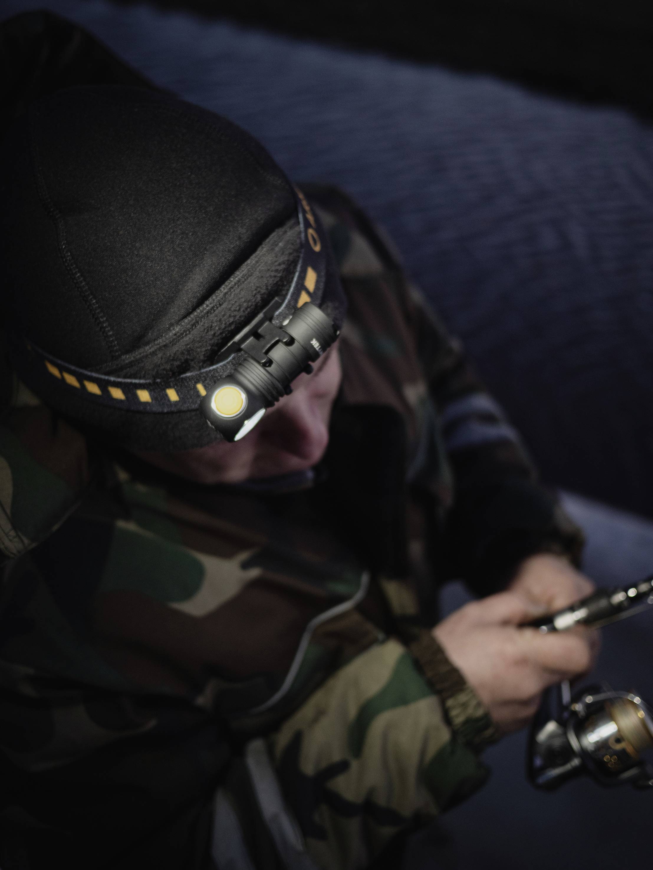 Osoba w ubraniu maskującym z czołówką łowi ryby, trzymając wędkę, jest w pełni skoncentrowana na pracy, na ciemnym tle w tle.