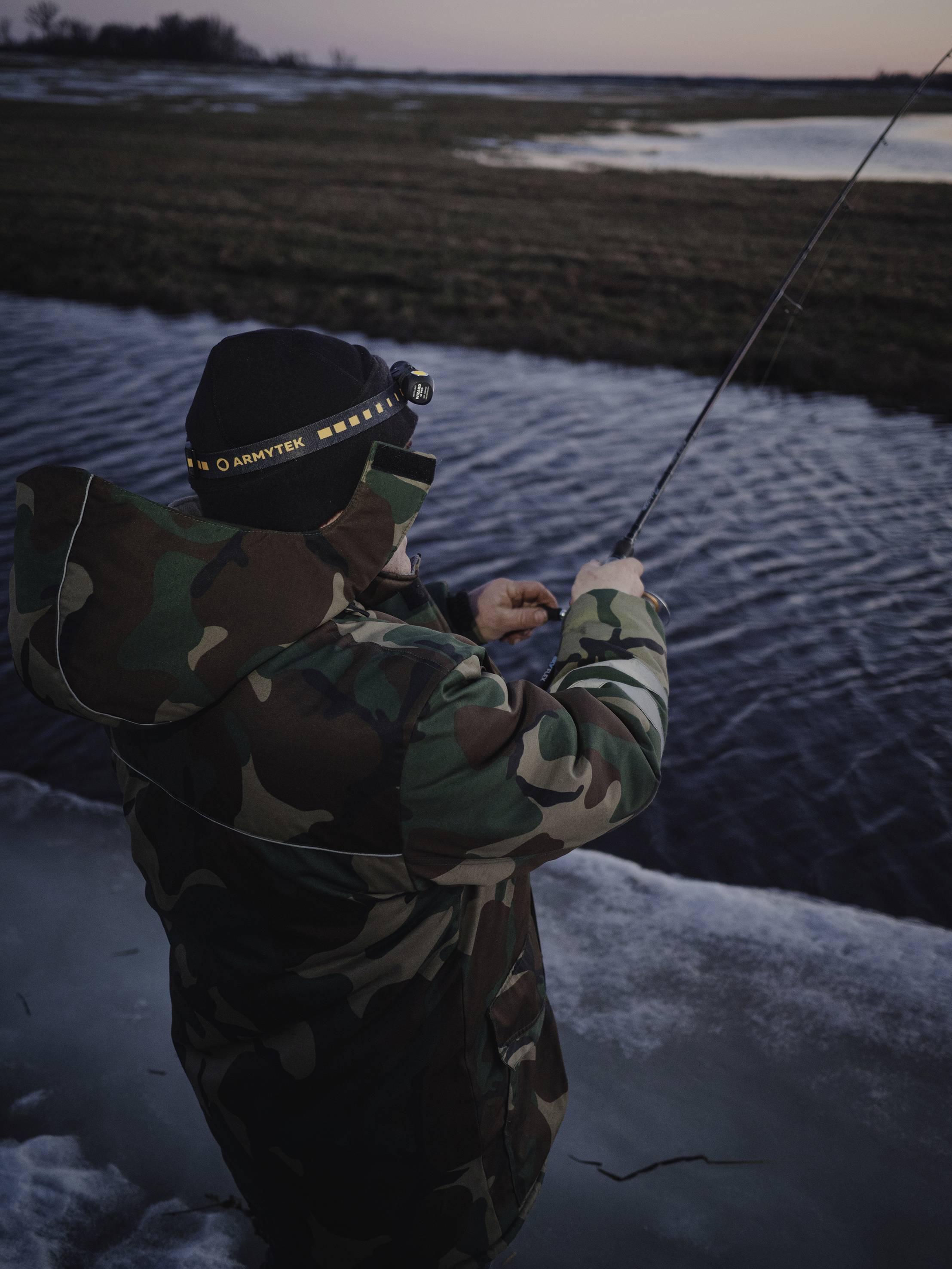 Osoba w kurtce maskującej stoi nad brzegiem rzeki, trzymając wędkę i patrząc w dal. Zapada zmierzch.