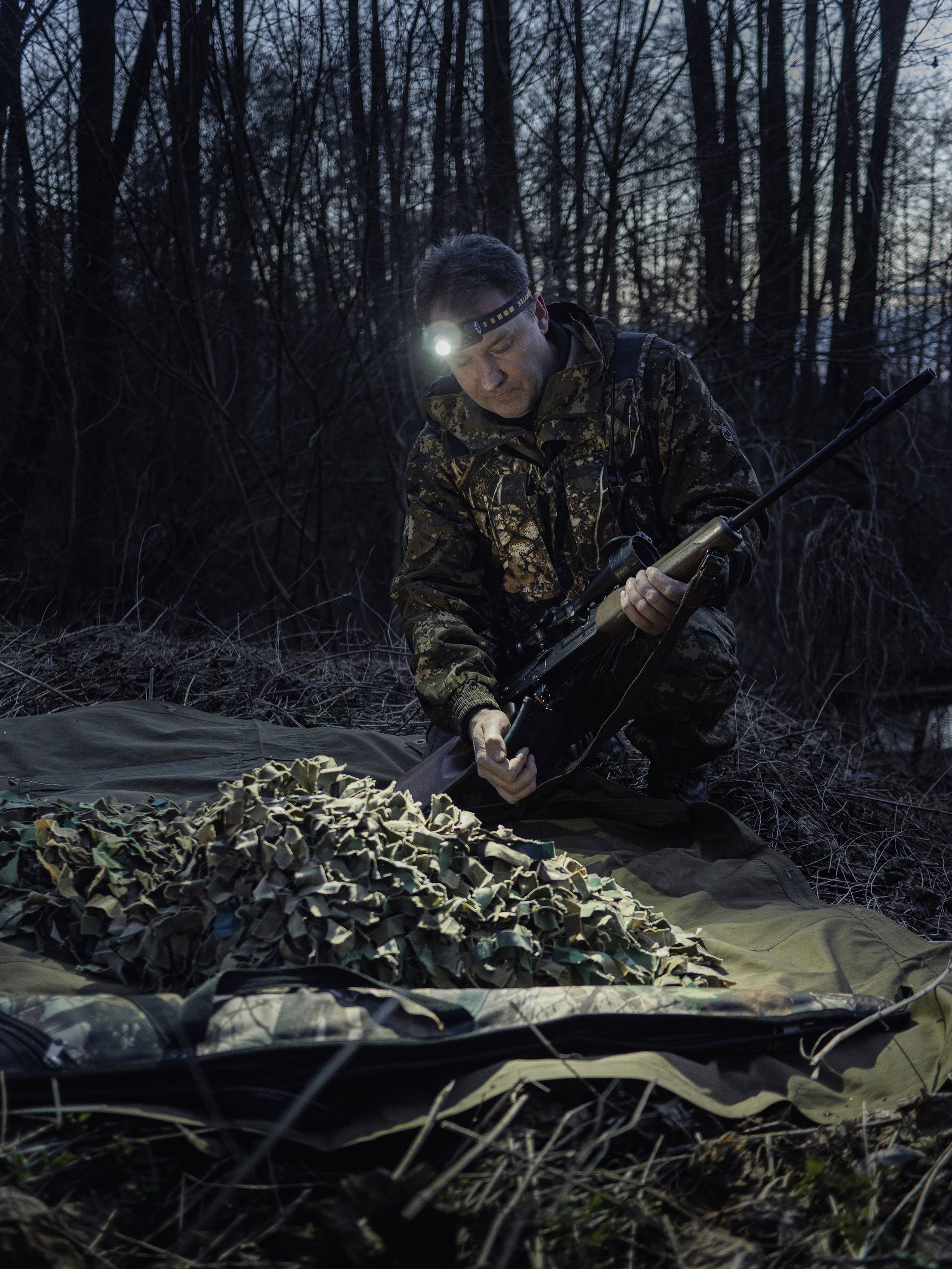 Mężczyzna w kamuflażu i czołówce klęczy obok stosu maskującego materiału w lesie o zmierzchu, trzymając w ręku broń.