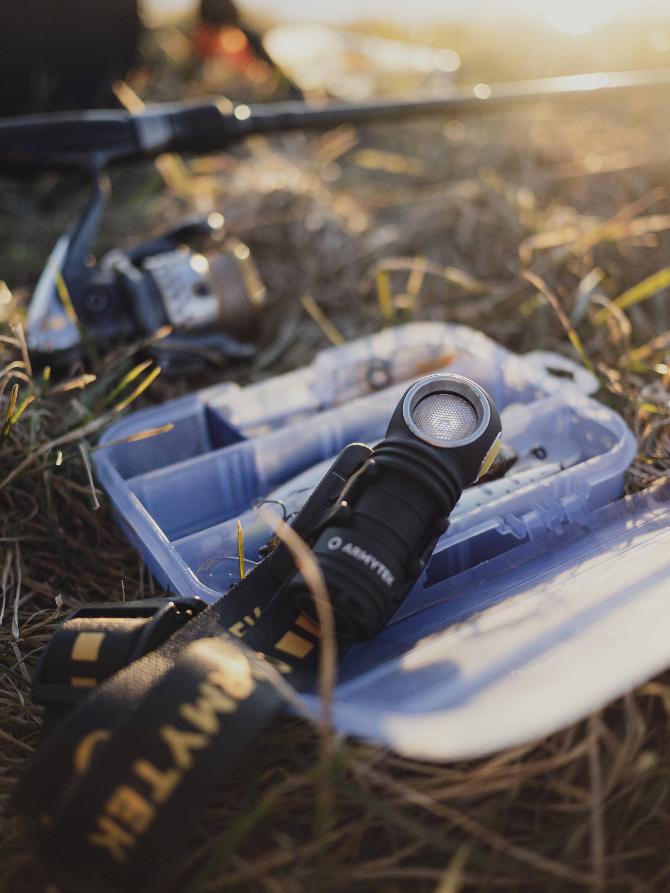 Torba wędkarska z wędkami i kołowrotkiem leży otwarta na trawie podczas zachodu słońca; latarka w pierwszym planie.