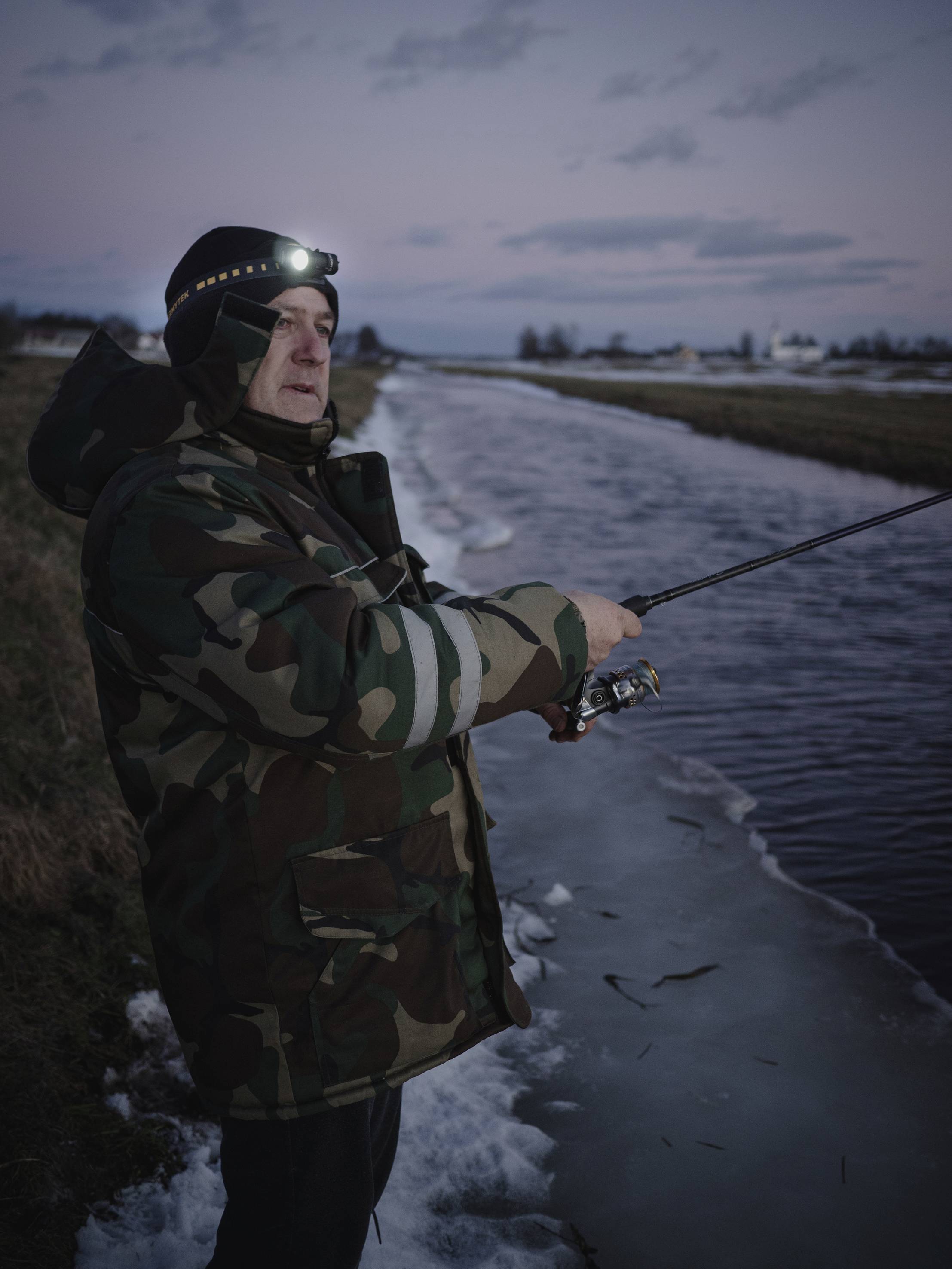 Osoba wędkuje nad zimowym brzegiem rzeki, ubrana w kurtkę maskującą i z czołówką. Na horyzoncie widoczny zmierzch.
