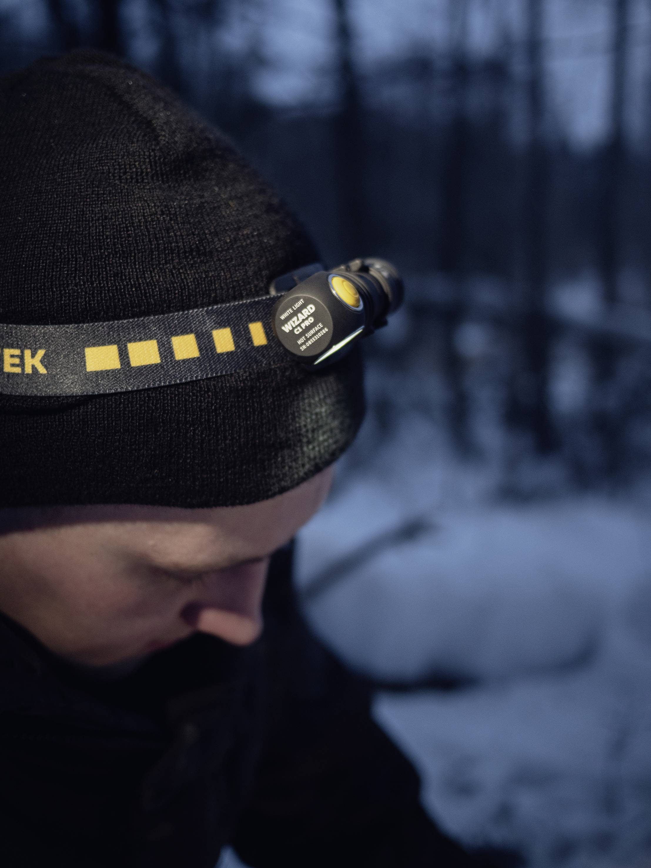 Osoba nosi czarną czapkę z czołówką, na dworze w zaśnieżonym krajobrazie leśnym o zmierzchu.