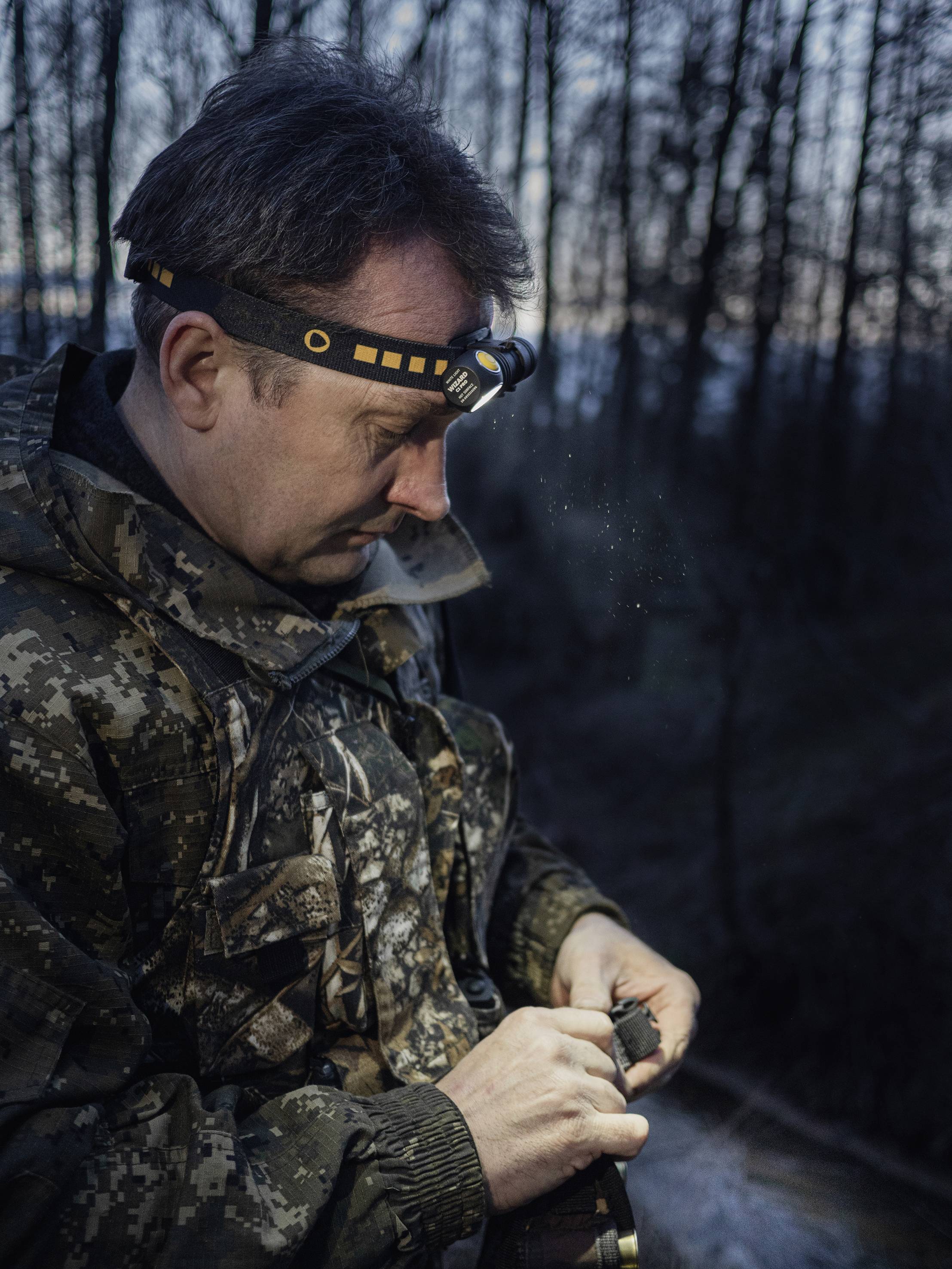 Mężczyzna w kamuflażu nosi czołówkę w ciemnym lesie. Koncentruje się na obiekcie trzymanym w rękach.