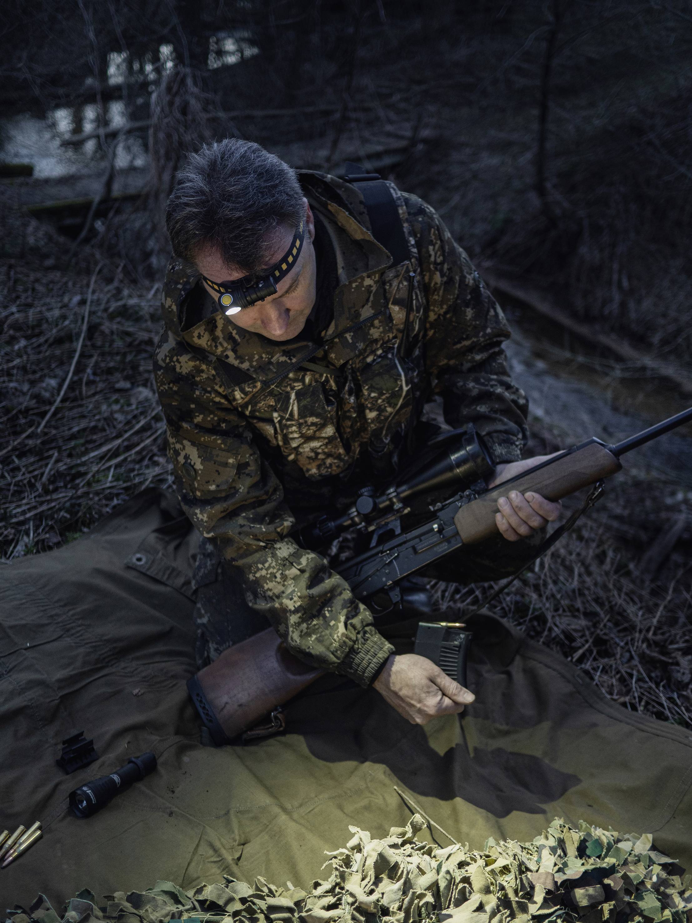 Osoba w stroju maskującym ładuje magazynek do broni w leśnym terenie nocą, wspierana światłem latarki.