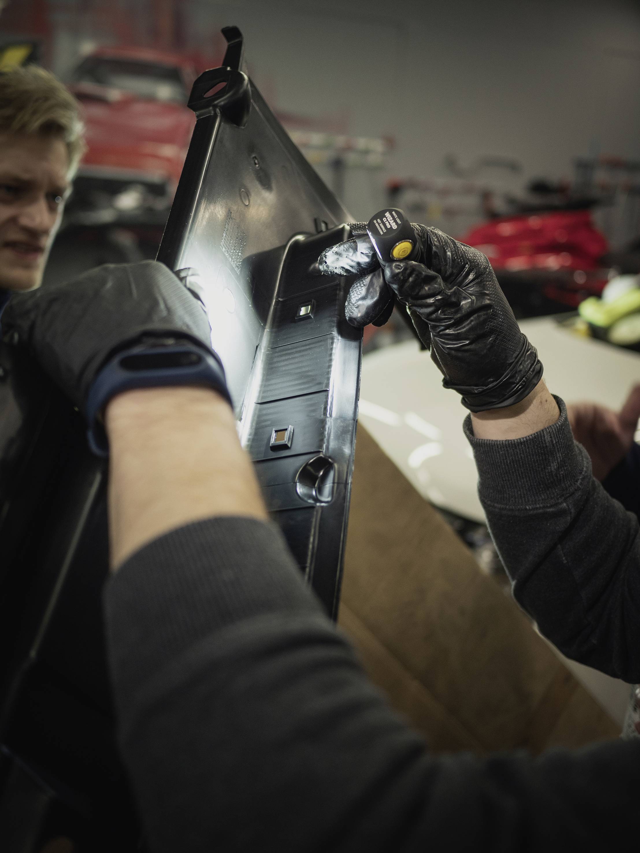 Dwie osoby wspólnie pracują nad częścią samochodową w warsztacie, jedna z nich trzyma latarkę i jest w rękawiczkach.