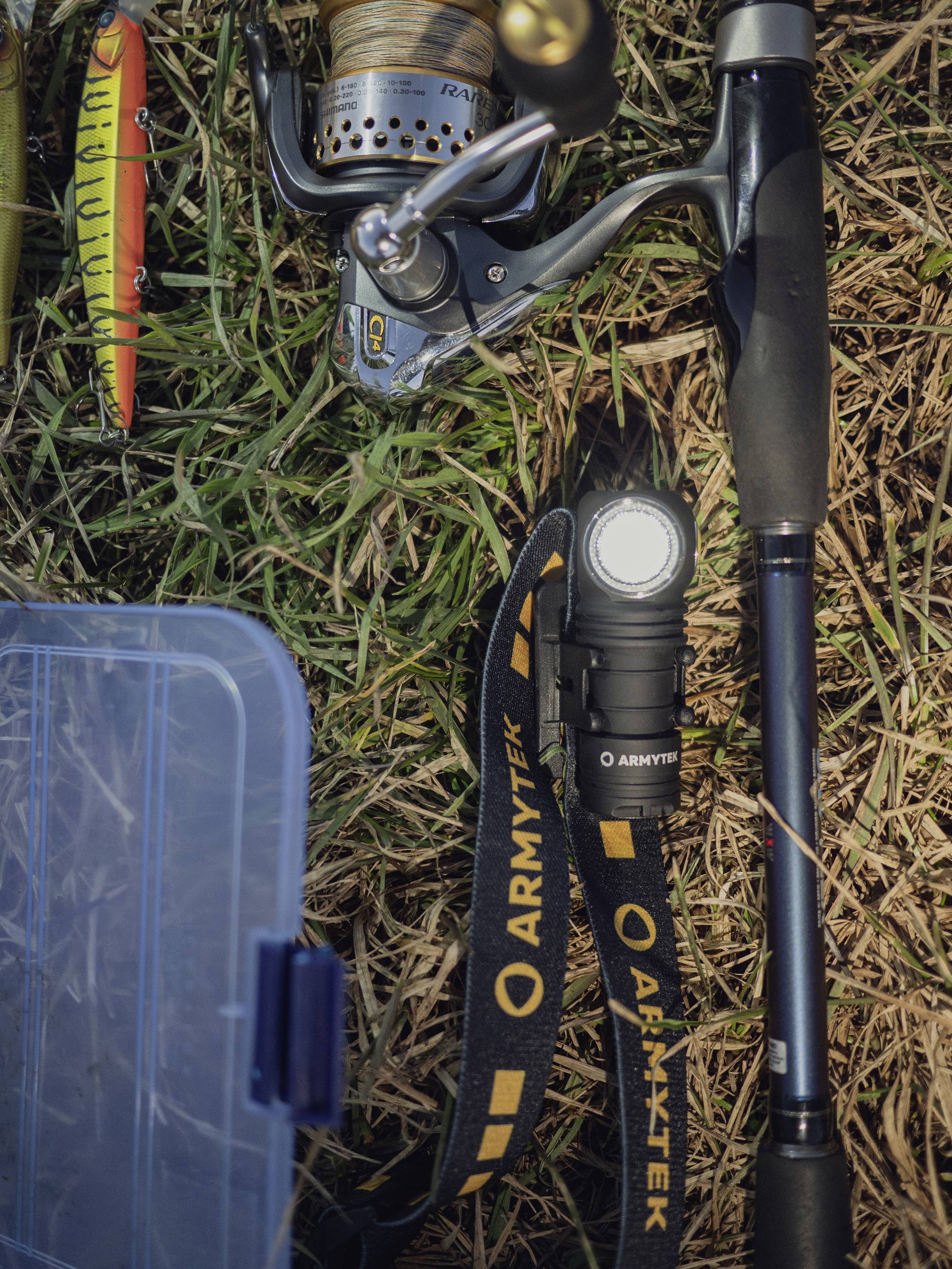 Akcesoria wędkarskie na trawie: Wędka i kołowrotek, przynęty, latarka z napisem 'ARMYTEK' oraz przezroczyste pudełko.