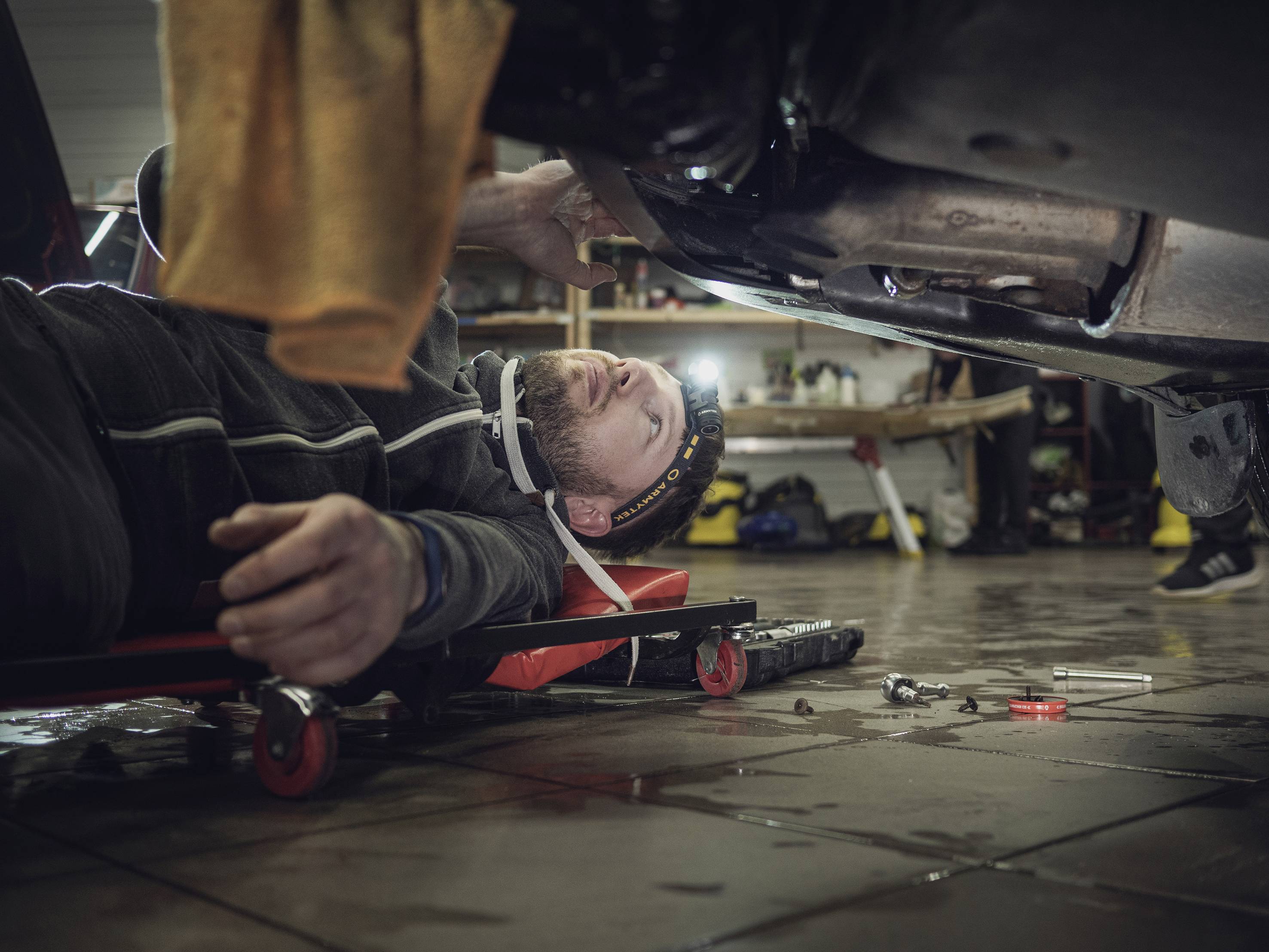 Mechanik leży na wózku warsztatowym pod samochodem w warsztacie i pracuje narzędziami przy dolnej części pojazdu.