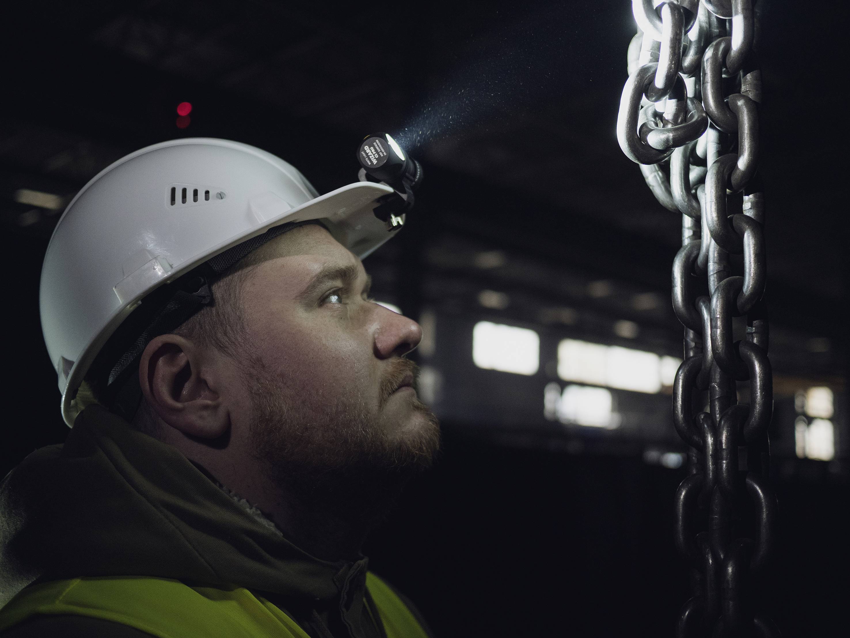 Robotnik w kasku i z czołówką dokładnie sprawdza ciężki łańcuch metalowy w ciemnym budynku przemysłowym.