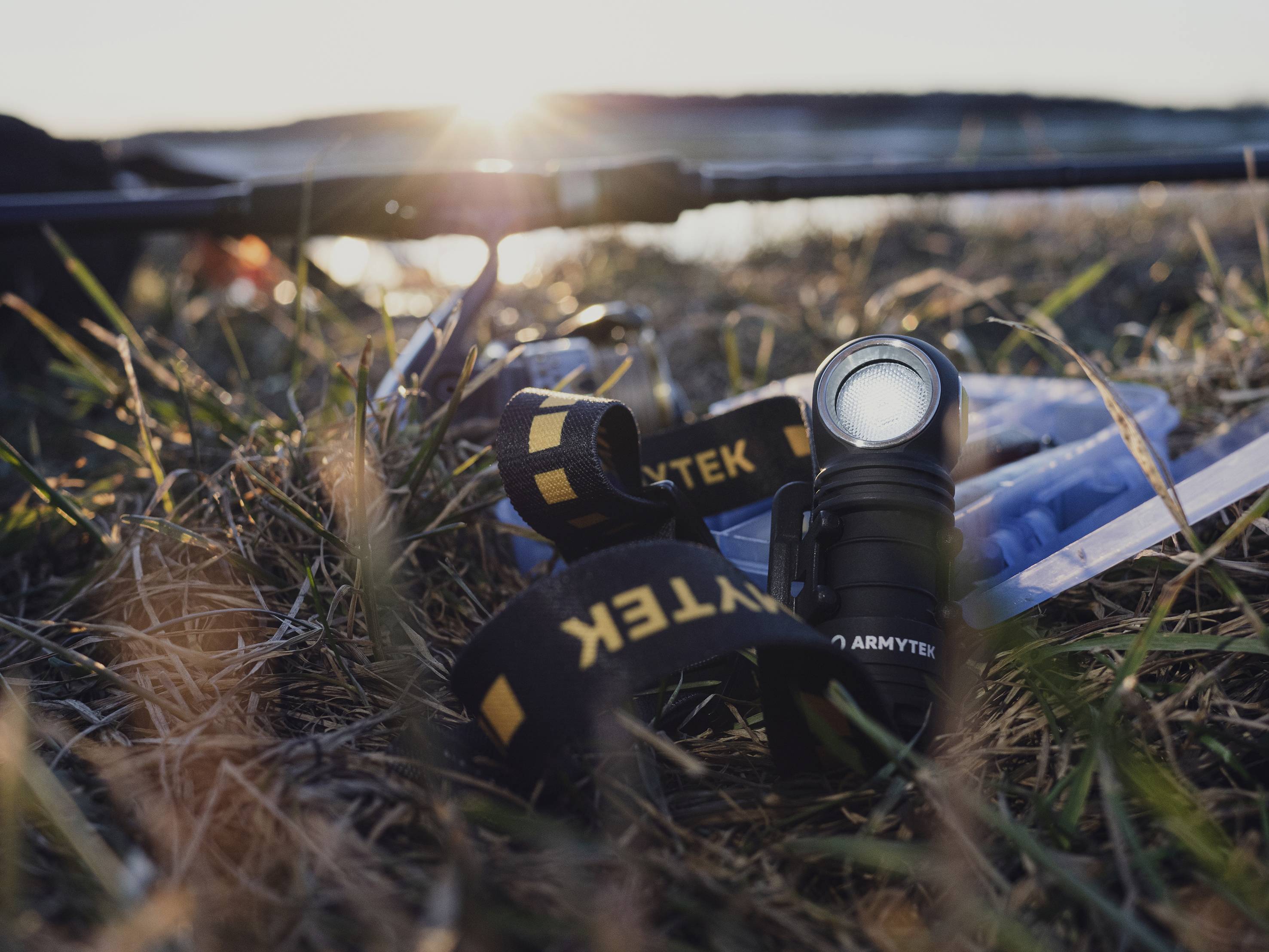 Latarka i akcesoria leżą na trawiastym gruncie podczas zachodu słońca, w pobliżu zbiornika wodnego. Klimatyczna scena plenerowa.