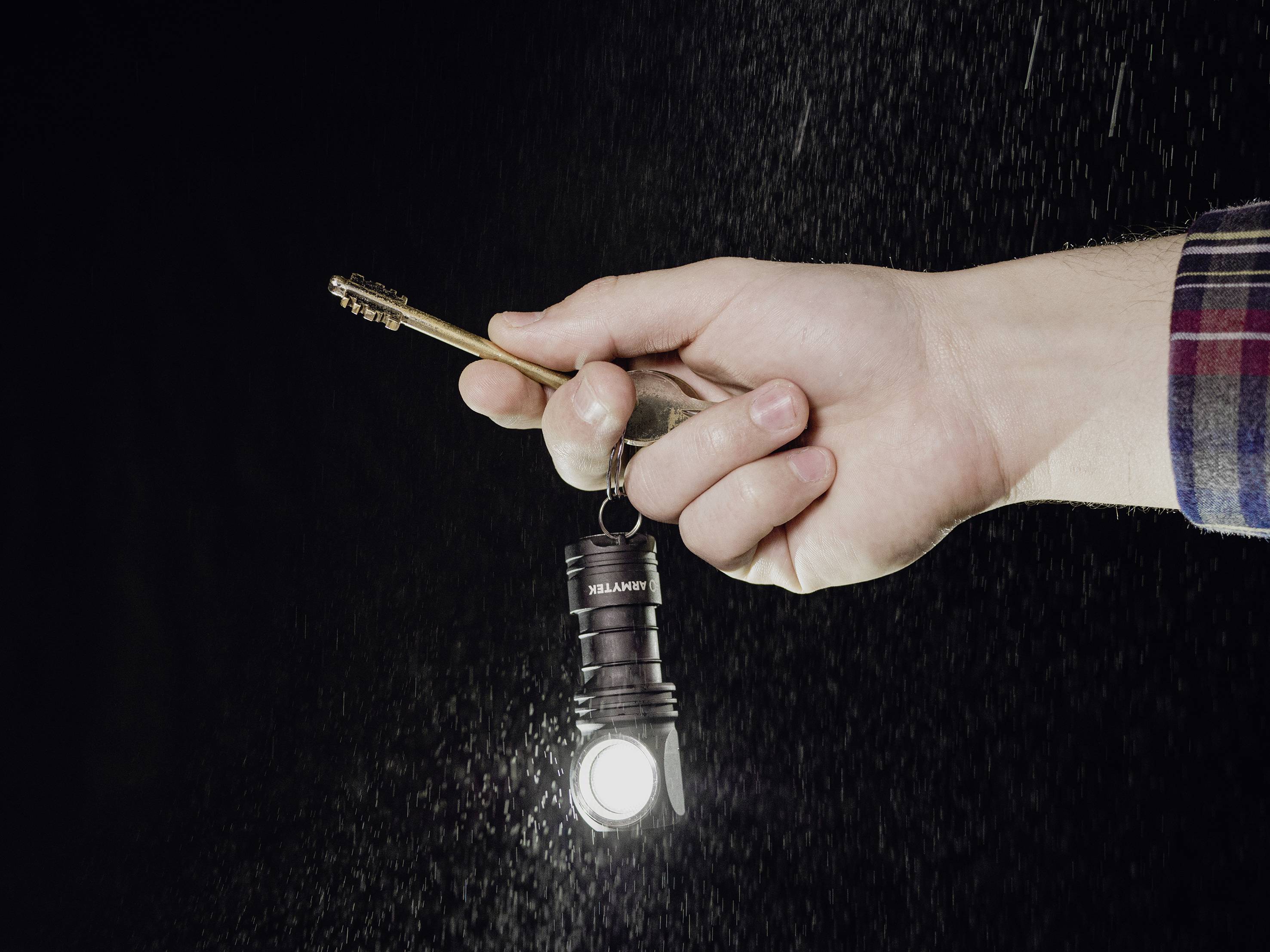 Jedna ręka trzyma klucz, do którego przymocowana jest mała latarka świecąca w ciemnym tle, prawdopodobnie podczas deszczu.