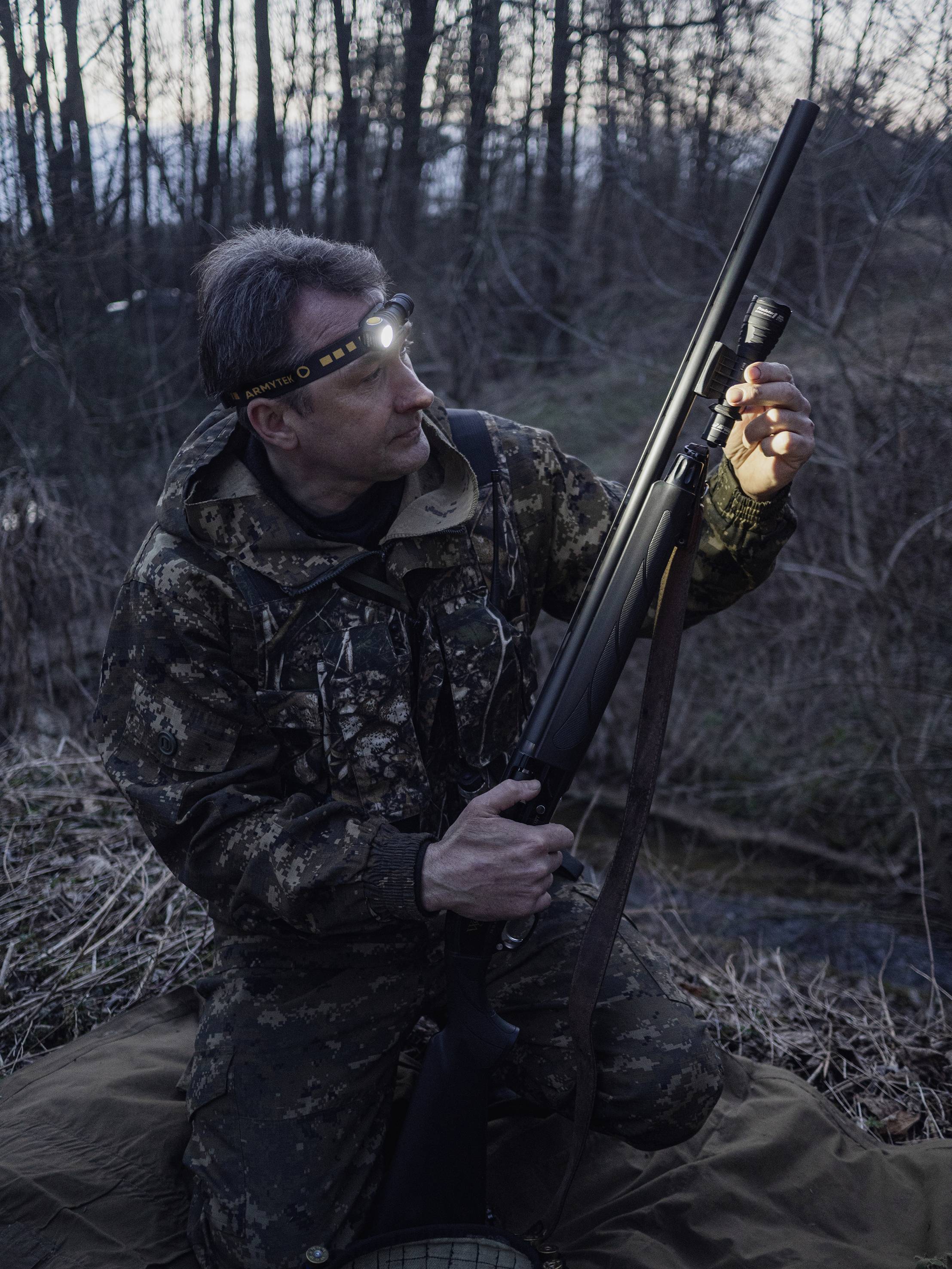Osoba w kamuflażu bada karabin w lesie o zmierzchu, nosząc czołówkę. Otoczenie jest zalesione i ciemne.