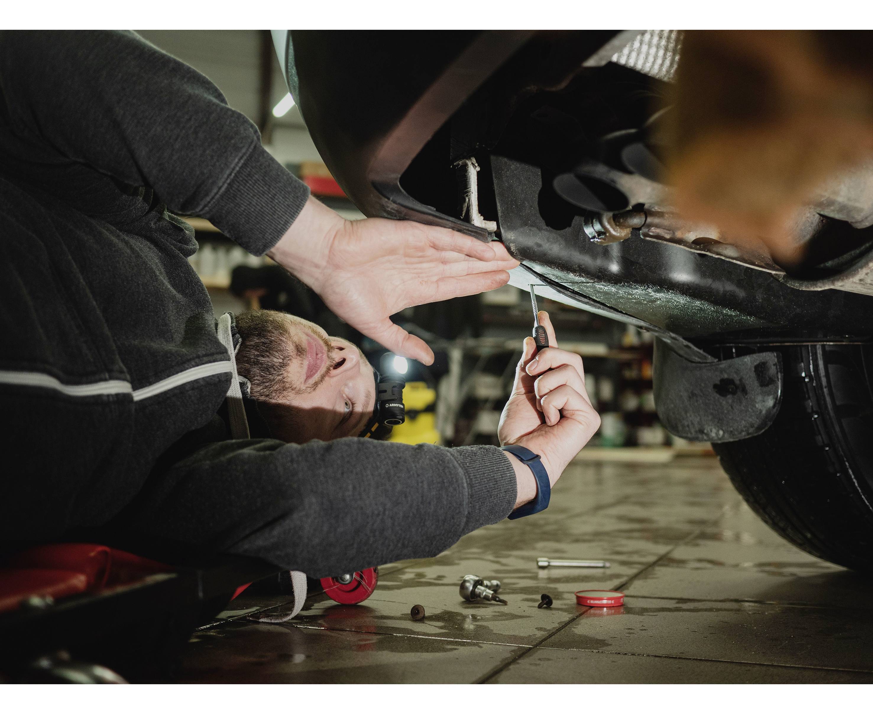 Osoba naprawia samochód, leżąc na plecach pod pojazdem i używając klucza do dokręcania śrub w warsztacie.