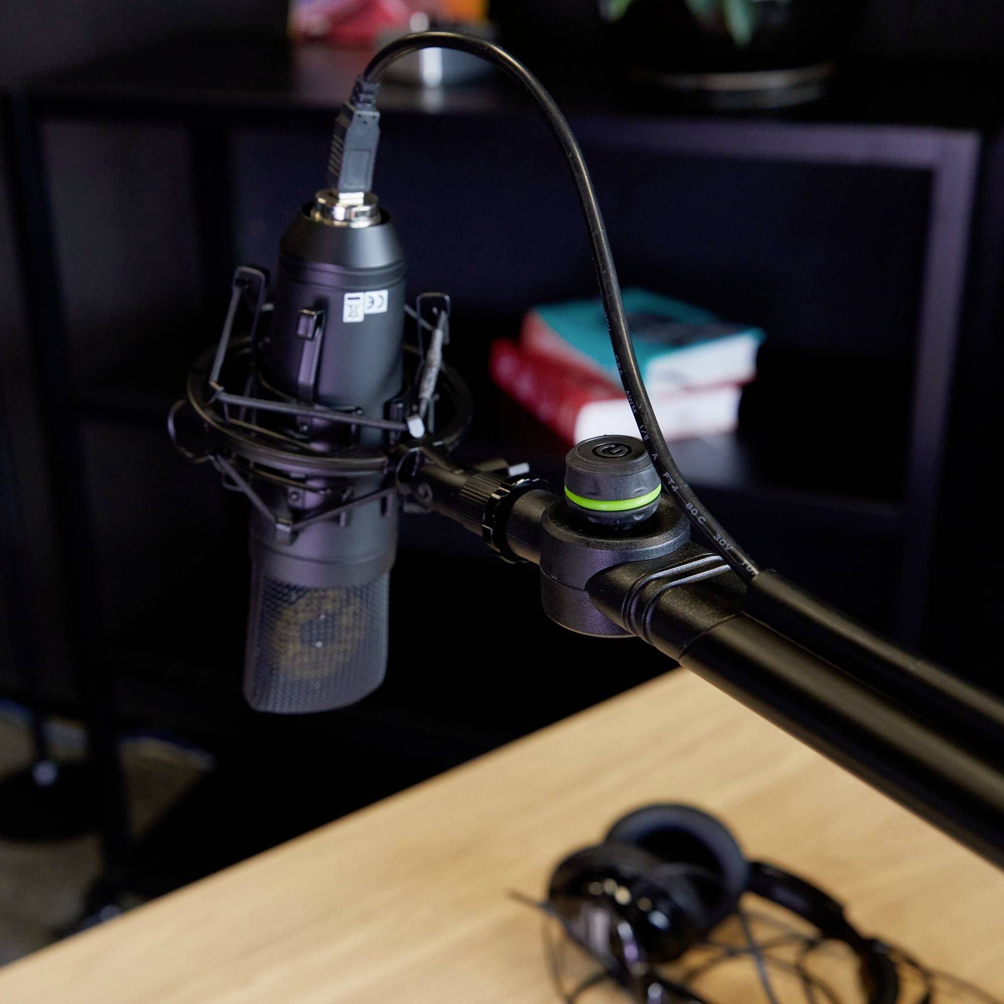 Mikrofon stojące na stole wskazuje na środowisko podcastowe lub nagraniowe. Obok leżą słuchawki, a w tle widoczne są książki.