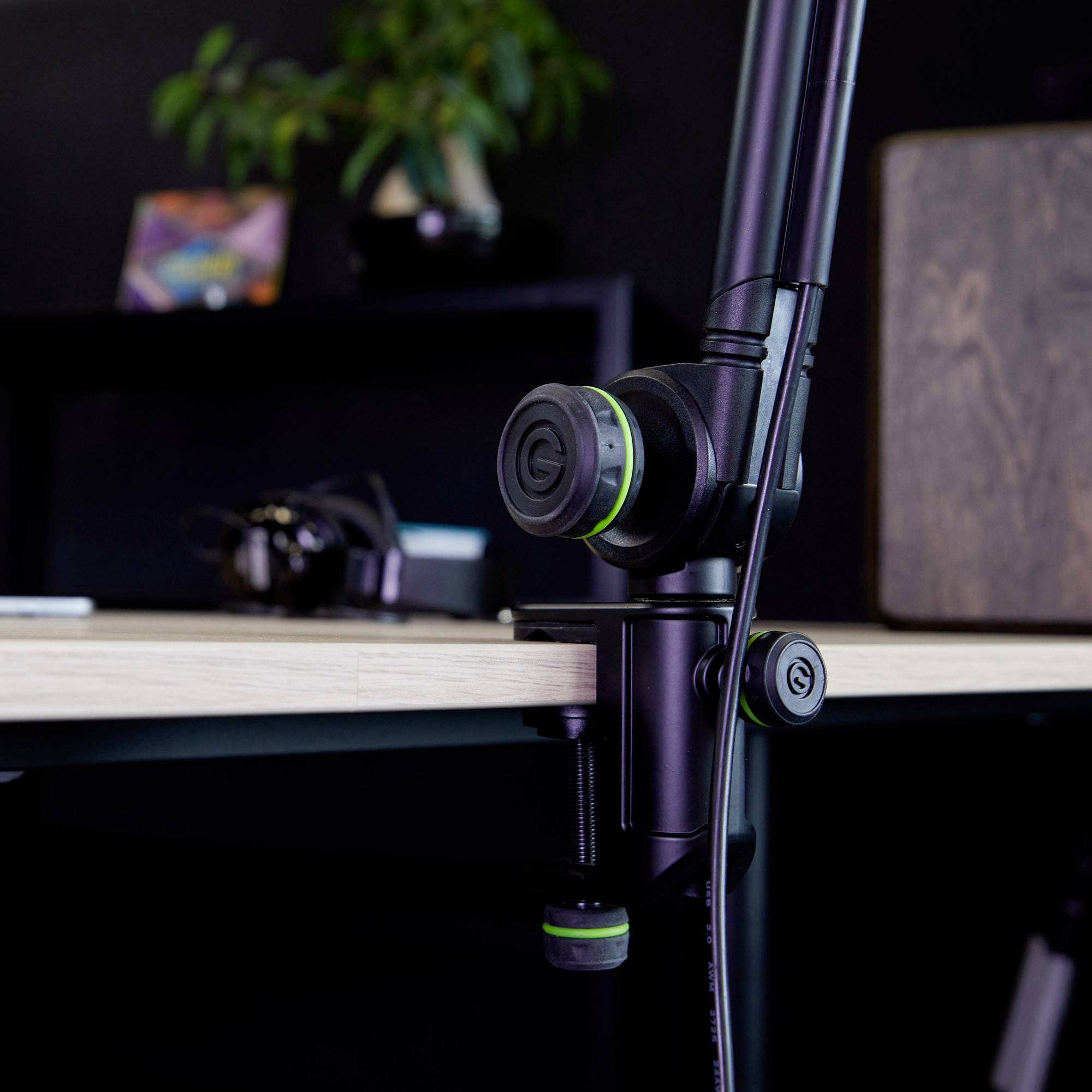 Ramię mikrofonu jest przymocowane do stołu. W tle, niewyraźnie widać słuchawki i roślinę.