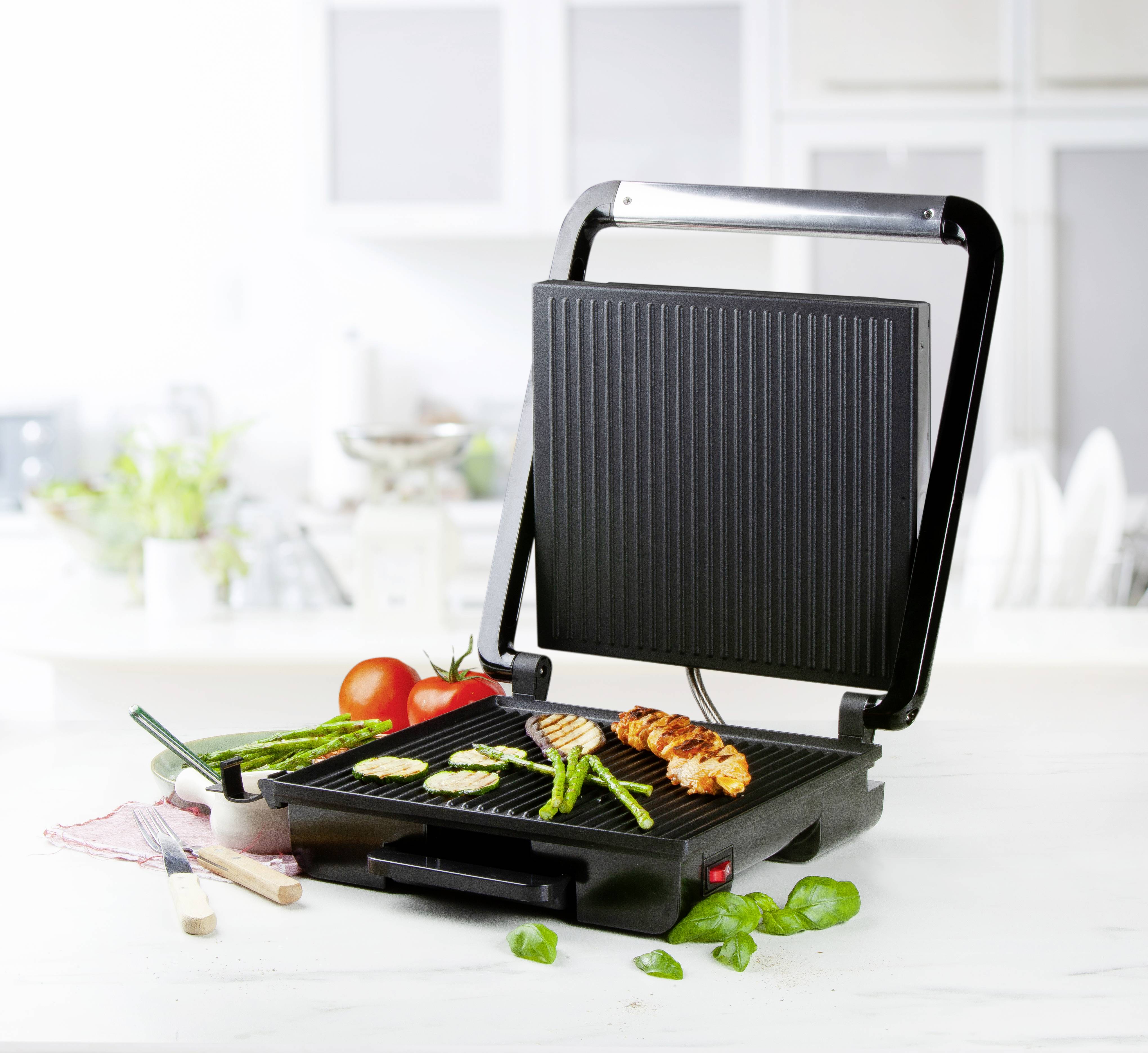 Elektryczny grill na stole kuchennym z grillowanymi warzywami i kurczakiem. W tle stoją przybory kuchenne.