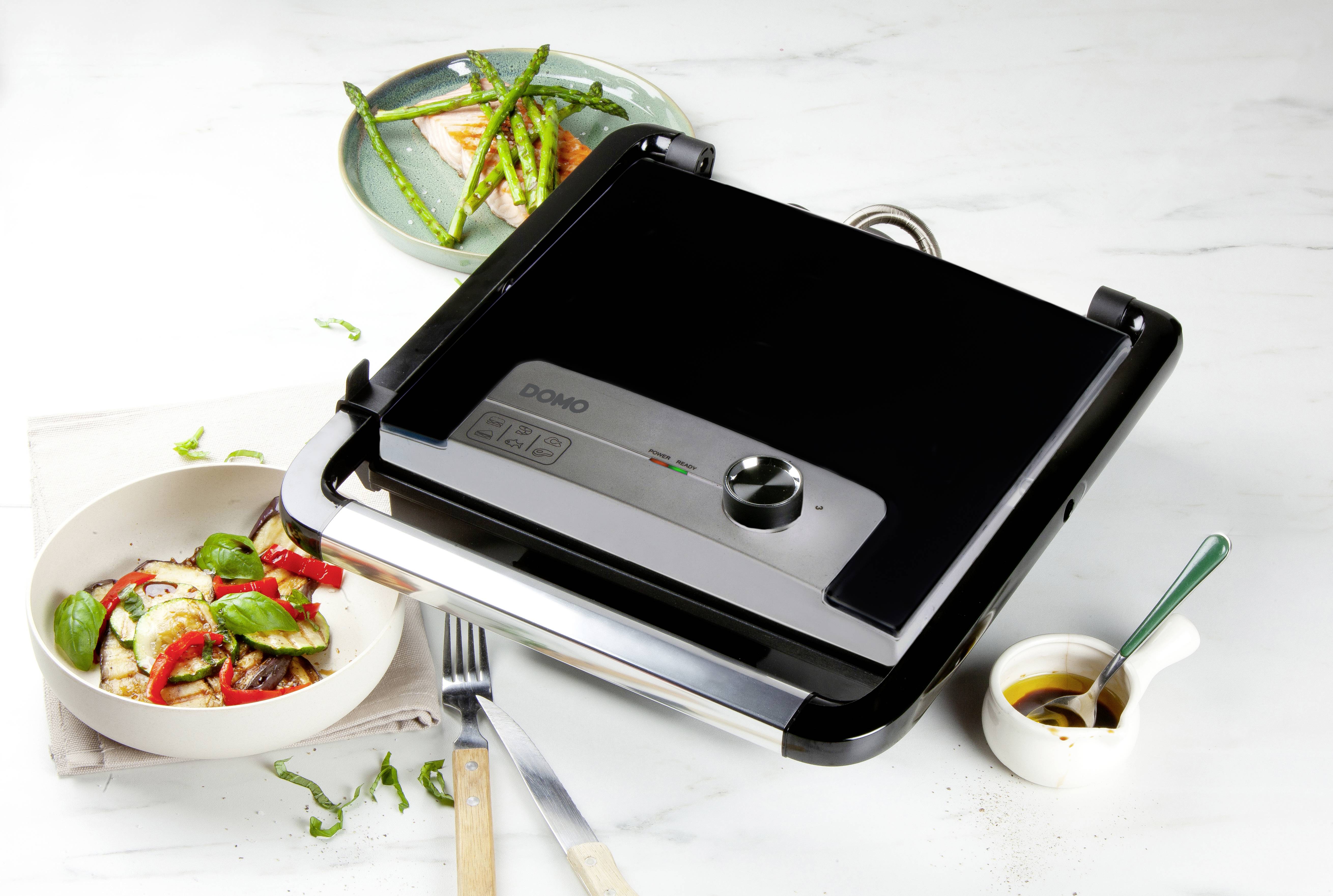 Elektryczny grill stołowy stojący na stole obok talerza z grillowanymi warzywami, sałatką i małą miseczką sosu.