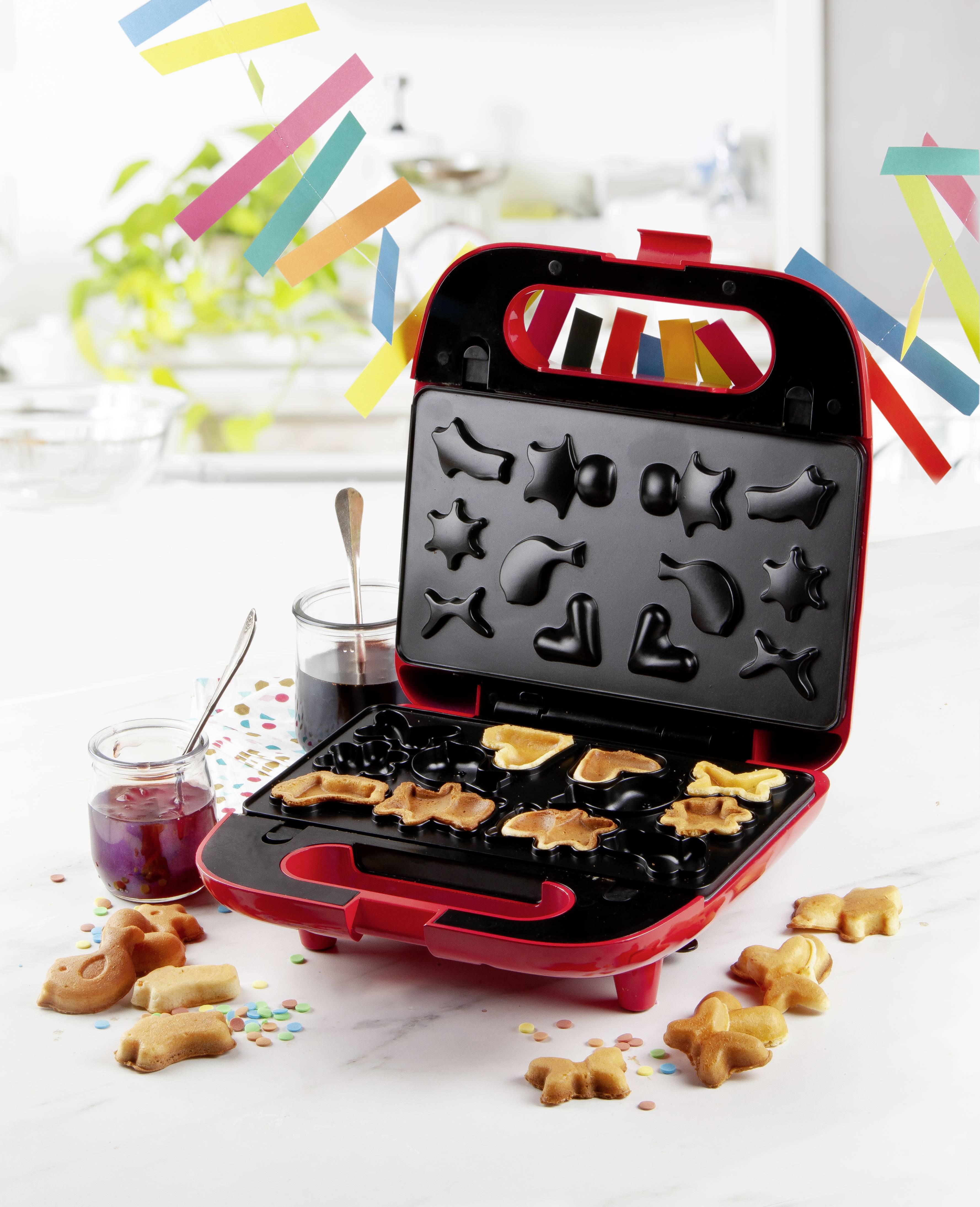 Czerwona gofrownica formuje gofry w różnych kształtach zwierząt i gwiazd. Na tle kolorowa dekoracja i słoiki z dżemem.