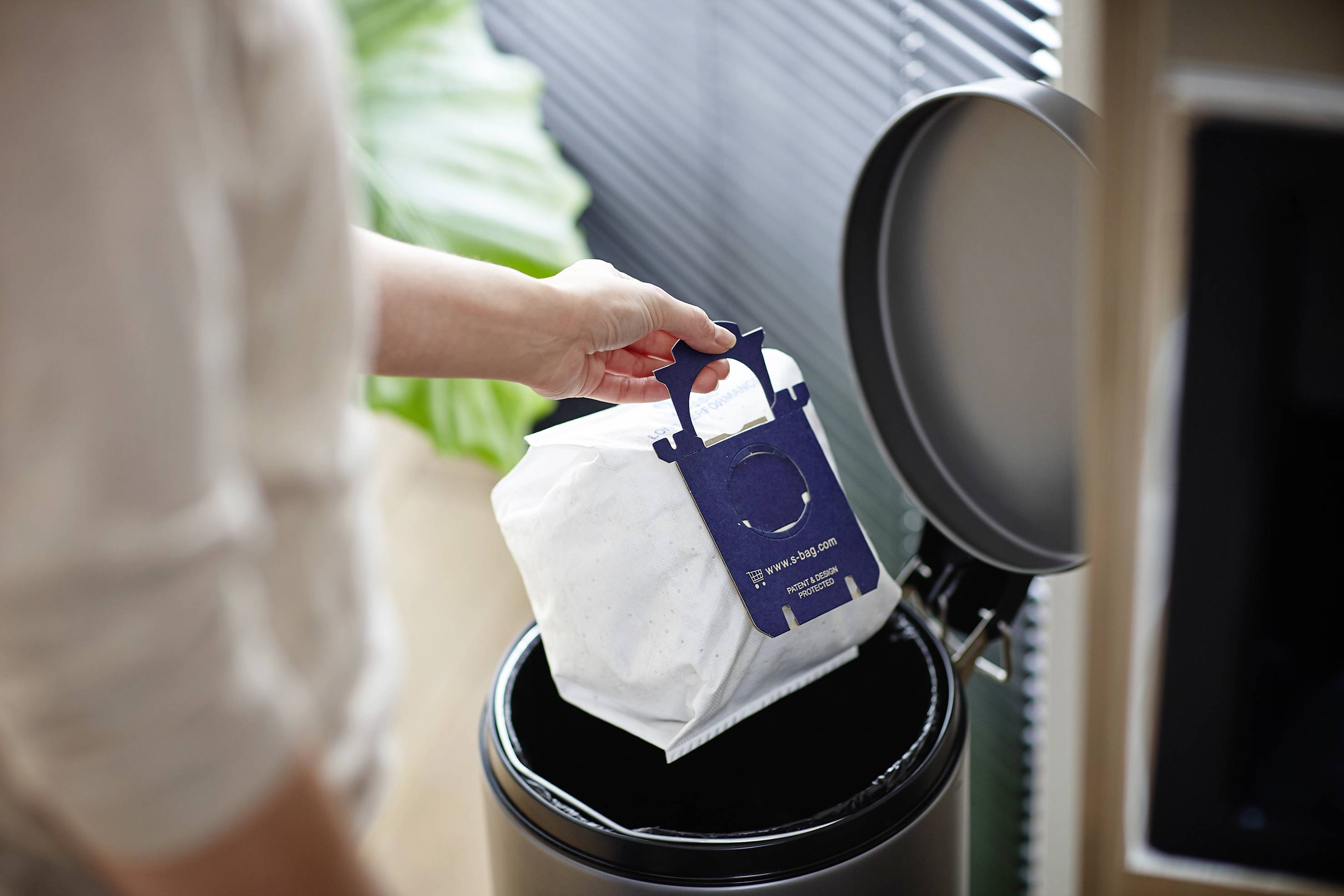 Osoba wyrzuca worek do odkurzacza do kosza na śmieci. W tle widać zieloną roślinę i żaluzje okienne.