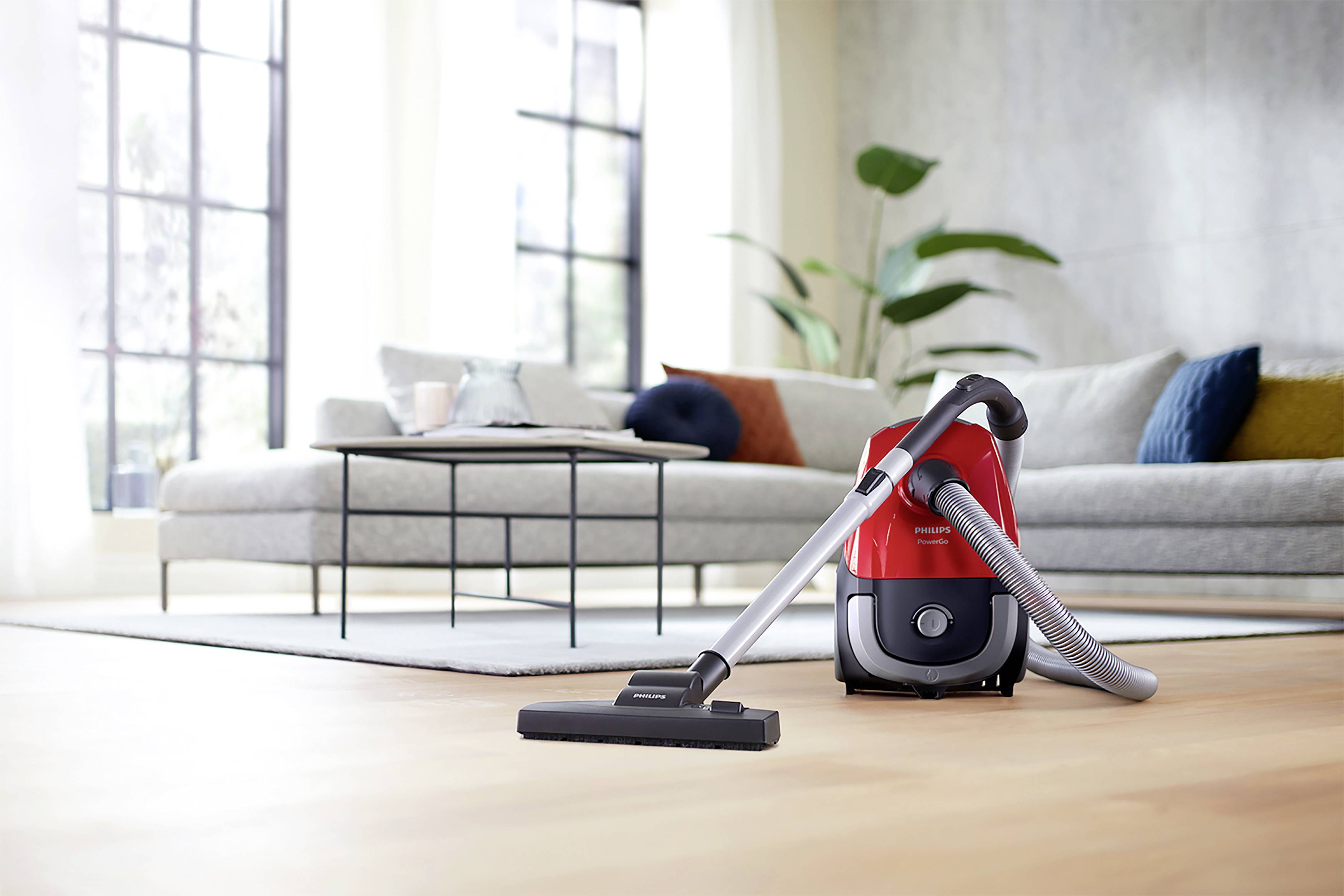 Czerwony odkurzacz stoi na jasnej drewnianej podłodze przed nowoczesnym salonem z kanapą, stolikiem kawowym i dużymi oknami.