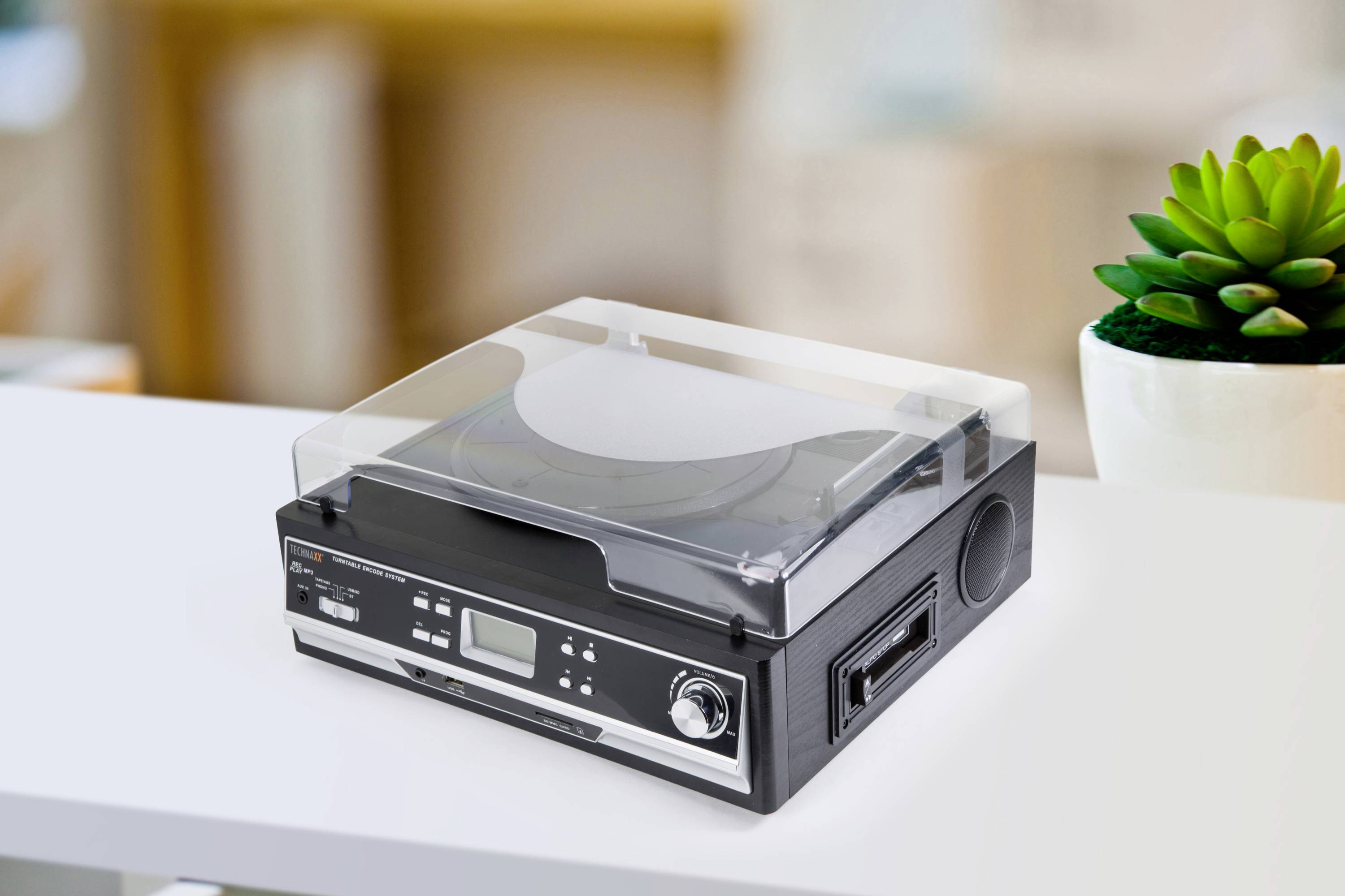 Gramofon z przezroczystą osłoną stoi na stole. W tle widać zieloną roślinę.