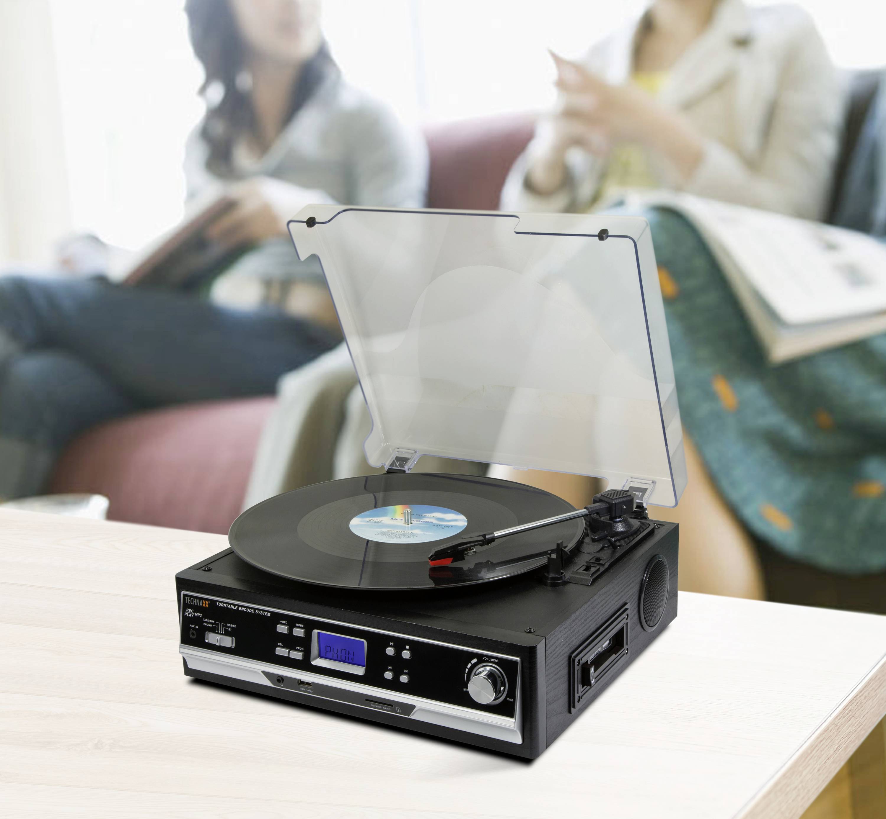 Nowoczesny gramofon z płytą winylową stoi na stole na pierwszym planie, a w tle, niewyraźnie, siedzą dwie osoby.