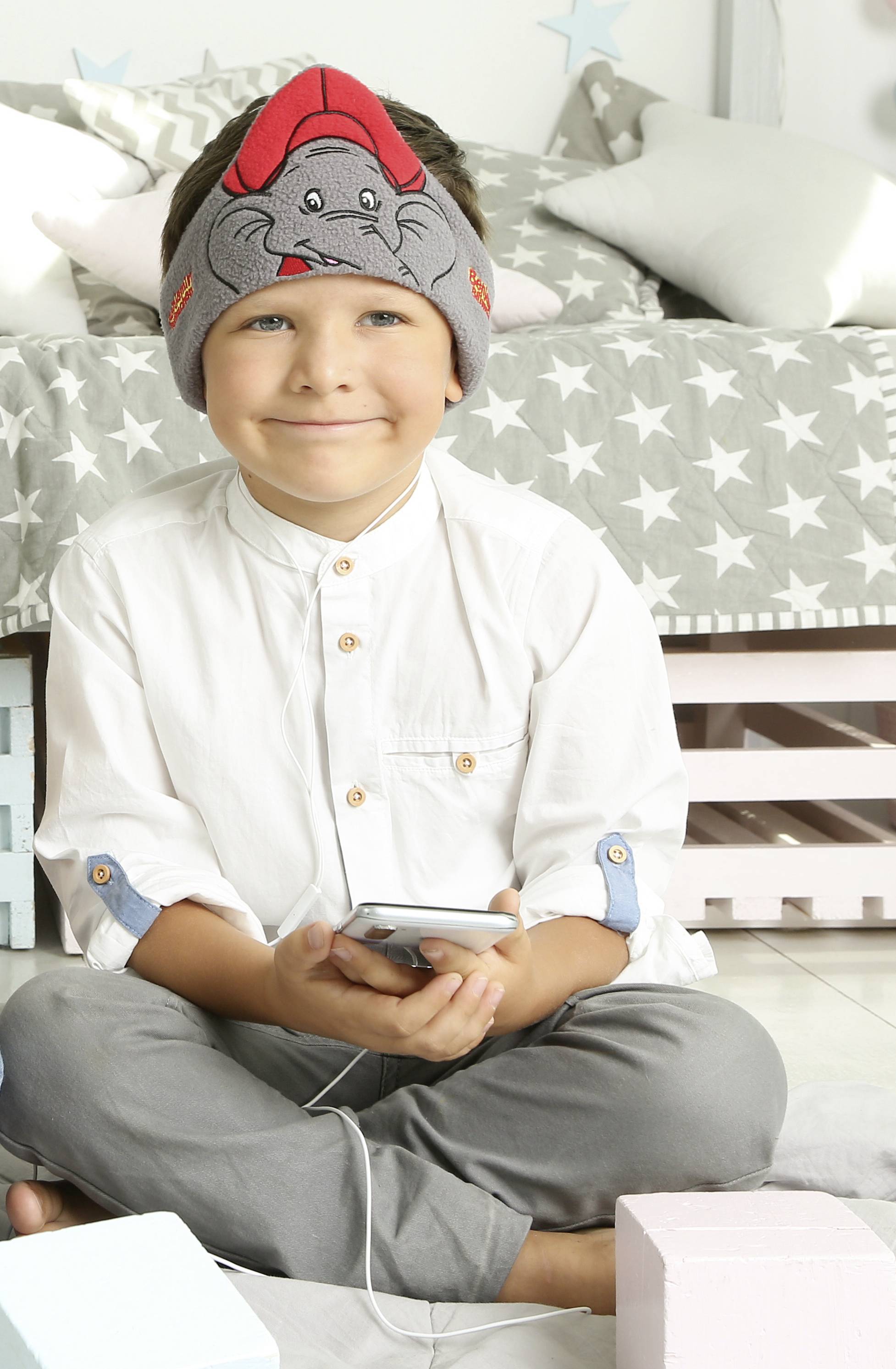 Uśmiechnięte dziecko w słuchawkach siedzi na podłodze i trzyma smartfon. W tle widać łóżko w gwiazdki.