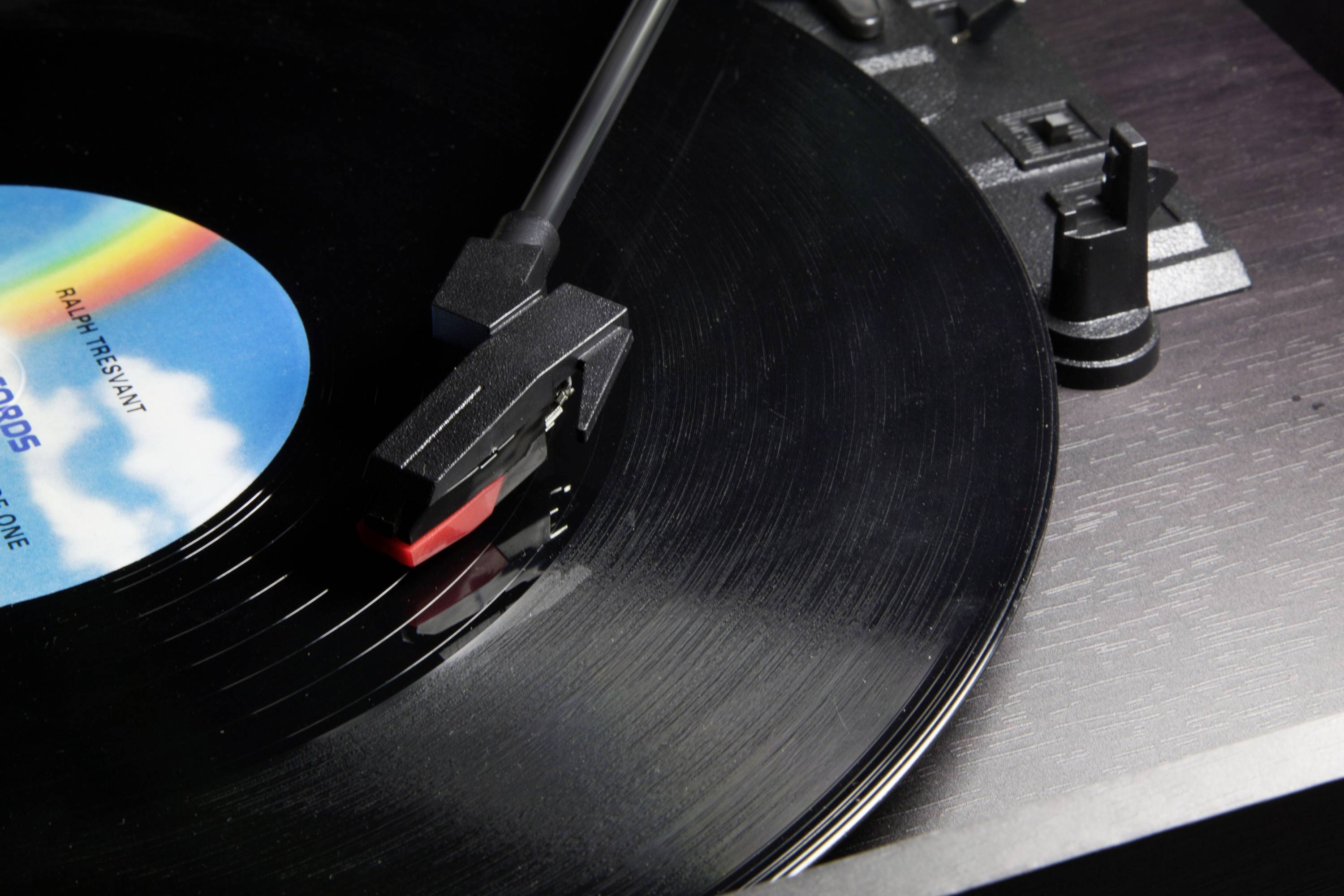 Gramofon z czarną płytą winylową. Ramiączka znajduje się na płycie. W tle widoczna jest etykieta w kolorach.