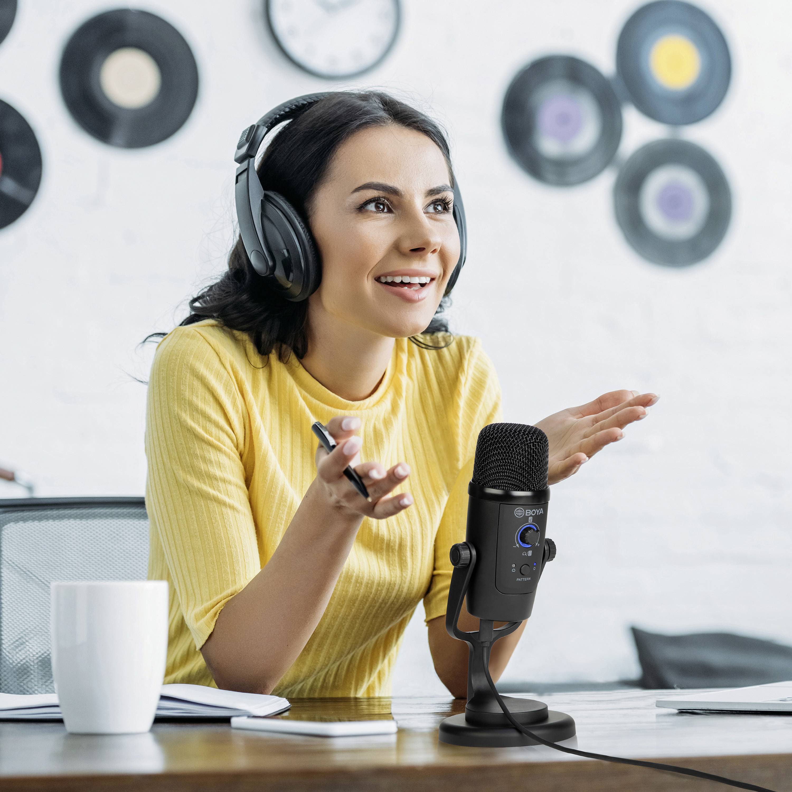 Kobieta w słuchawkach siedzi przy stole przed mikrofonem, wygląda na zaangażowaną. Na tle ściany widać płyty winylowe.