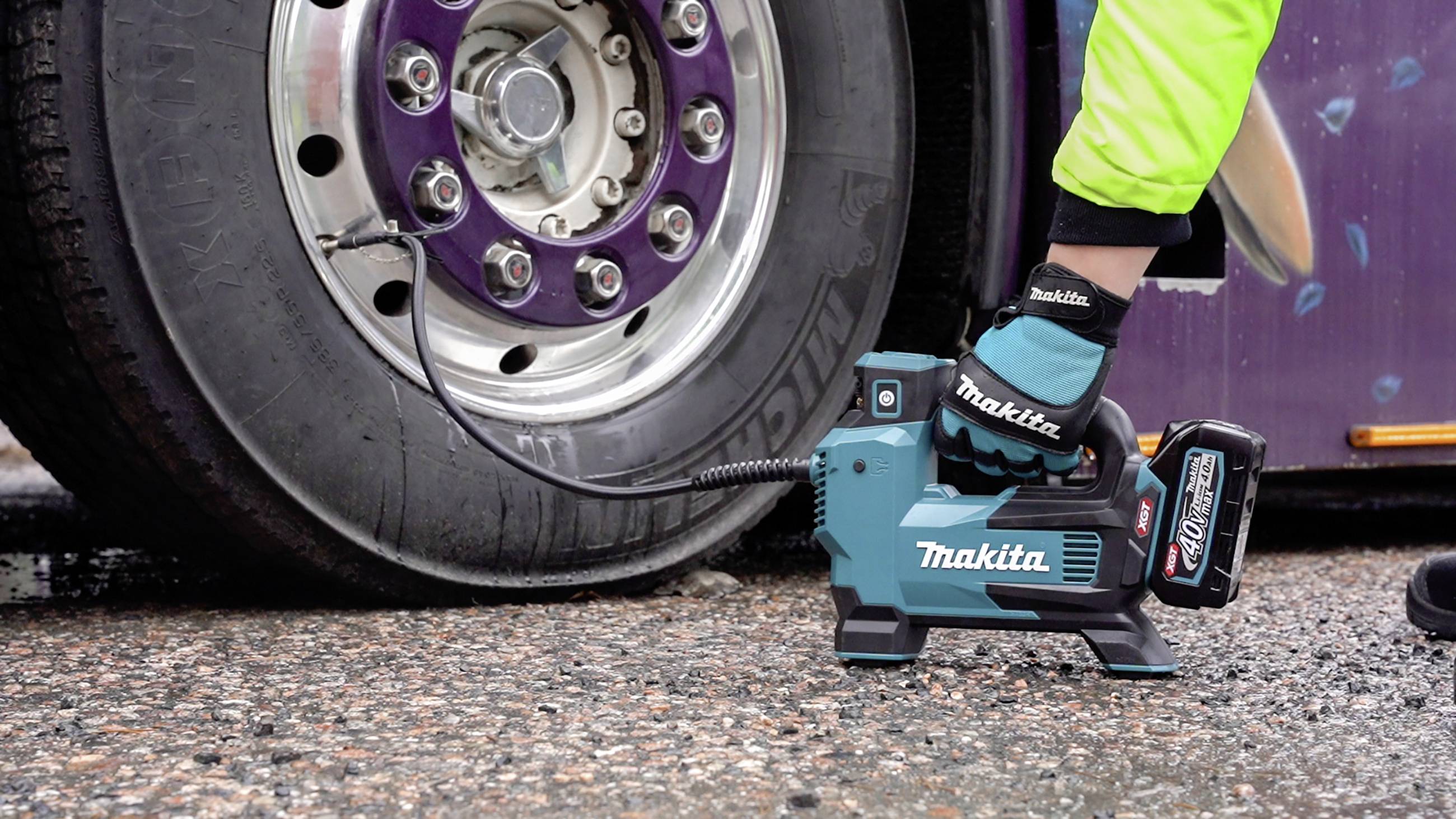 Akumulatorowy kompresor Makita pompuje opony ciężarówki. Jedna ręka trzyma urządzenie, które stoi na żwirowej powierzchni.
