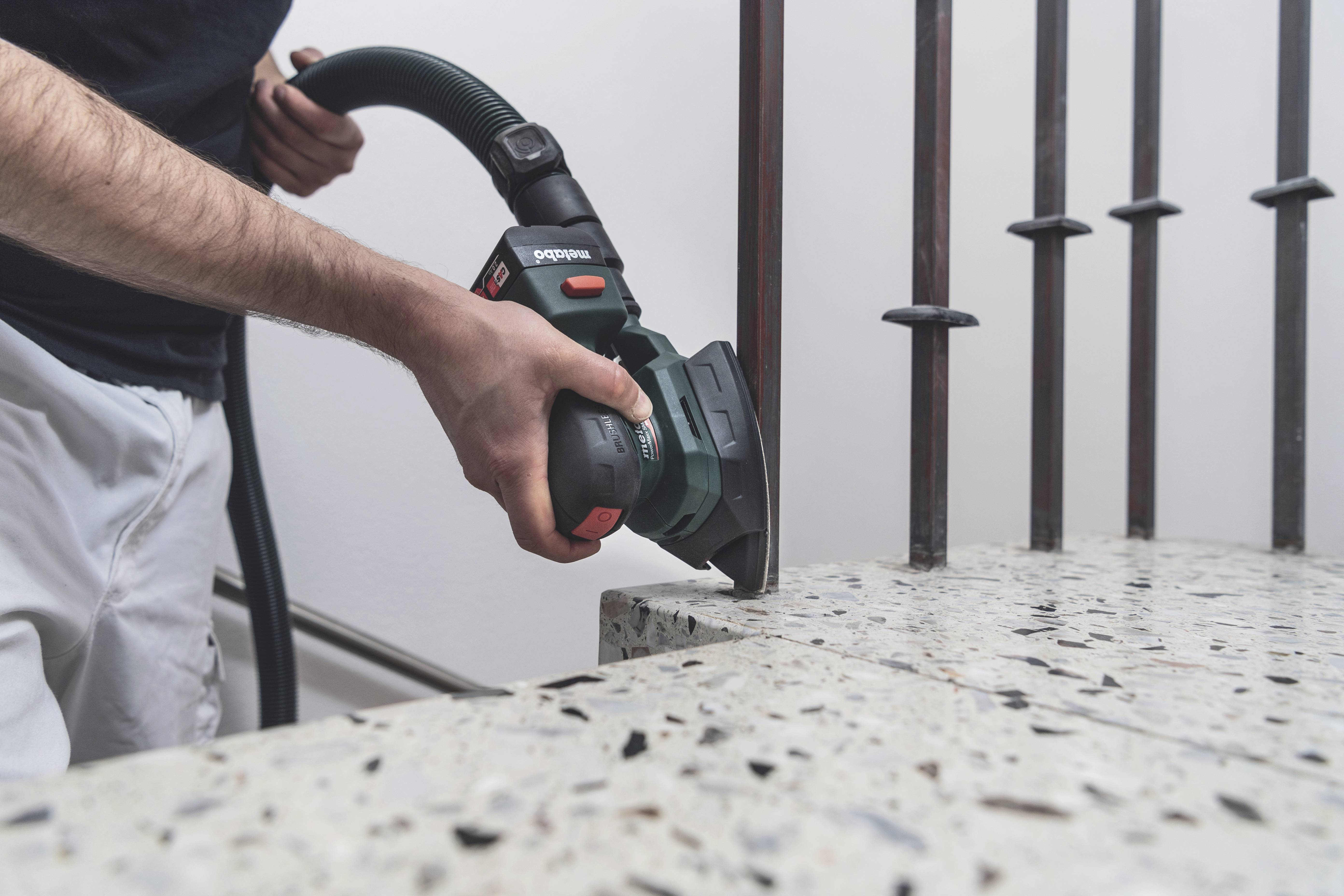 Osoba szlifuje krawędź bloku betonowego lub kamiennego elektronarzędziem. W tle widoczne są pręty metalowe.