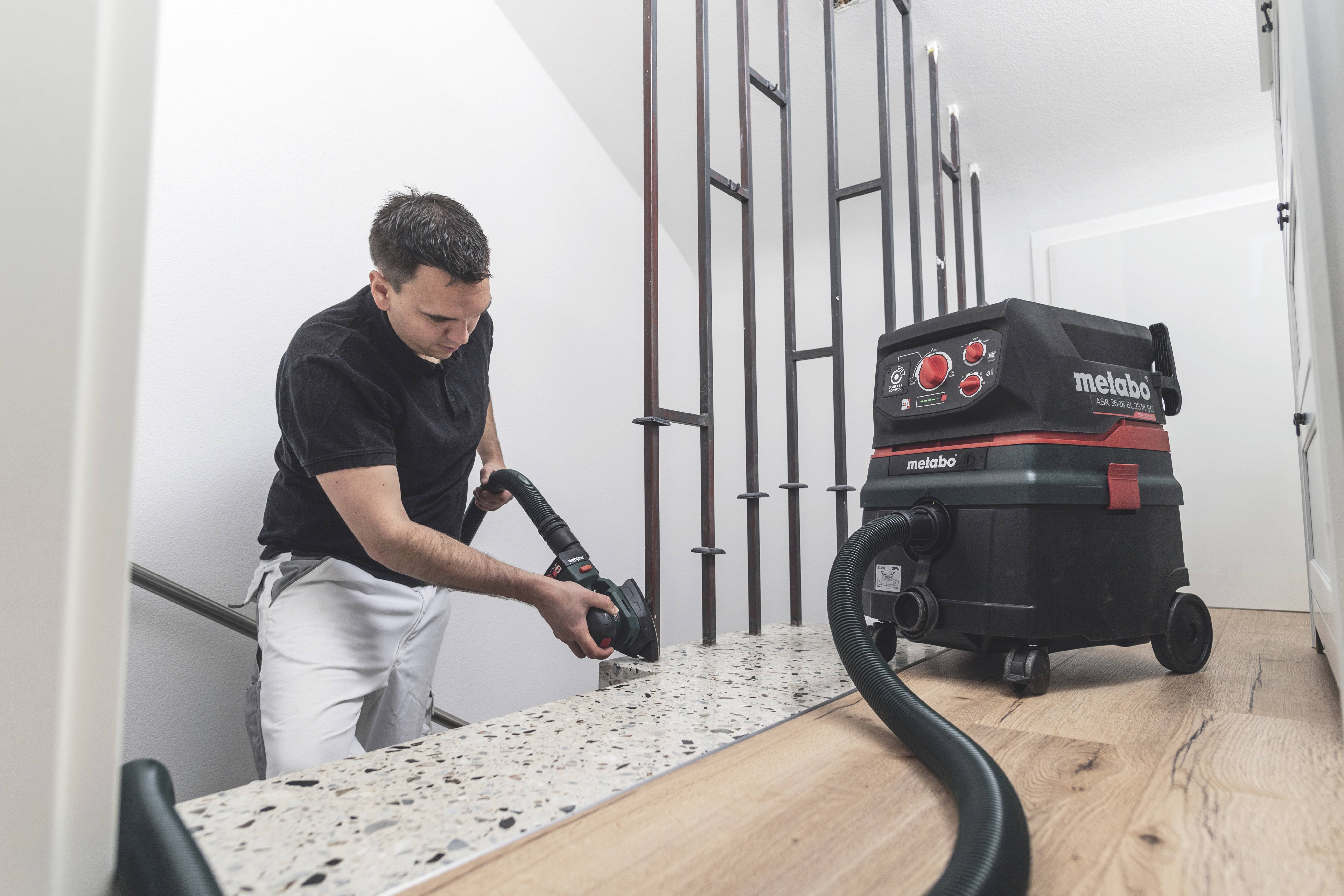 Mężczyzna używa przemysłowego odkurzacza do czyszczenia spocznika schodów. Odkurzacz jest czarny z czerwonymi akcentami.