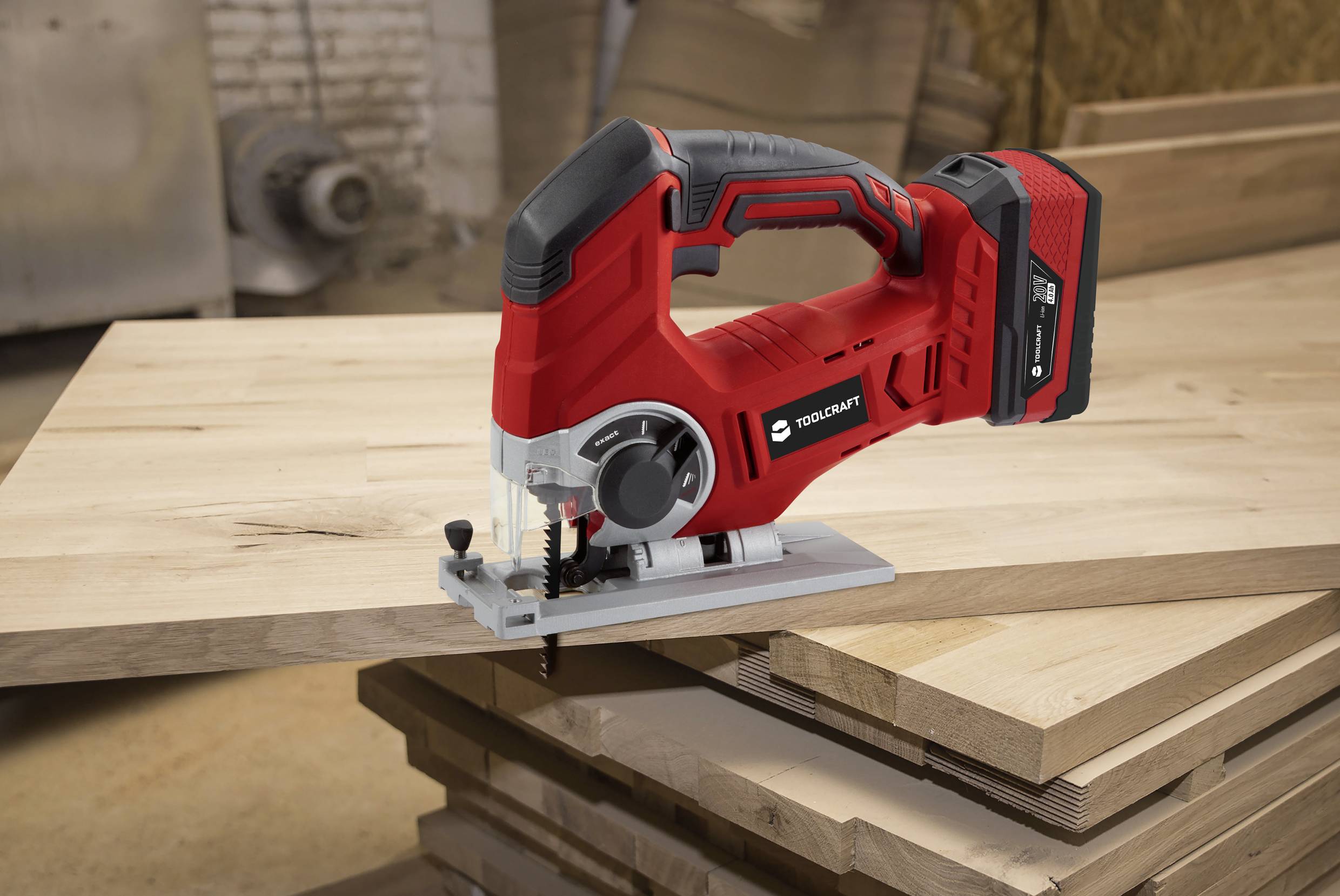 Czerwona akumulatorowa wyrzynarka na desce drewnianej w warsztacie. W tle widoczne są stosy drewna i nieostre narzędzia.